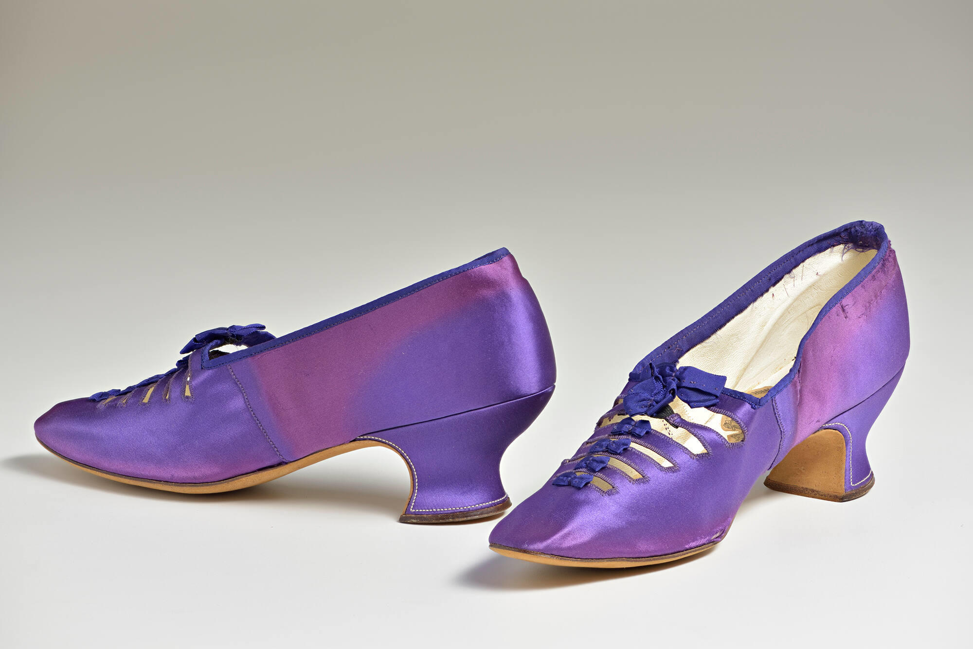 A pair of purple silk shoes with a tapered low heel and six straps across the vamp with purple grosgrain bows.