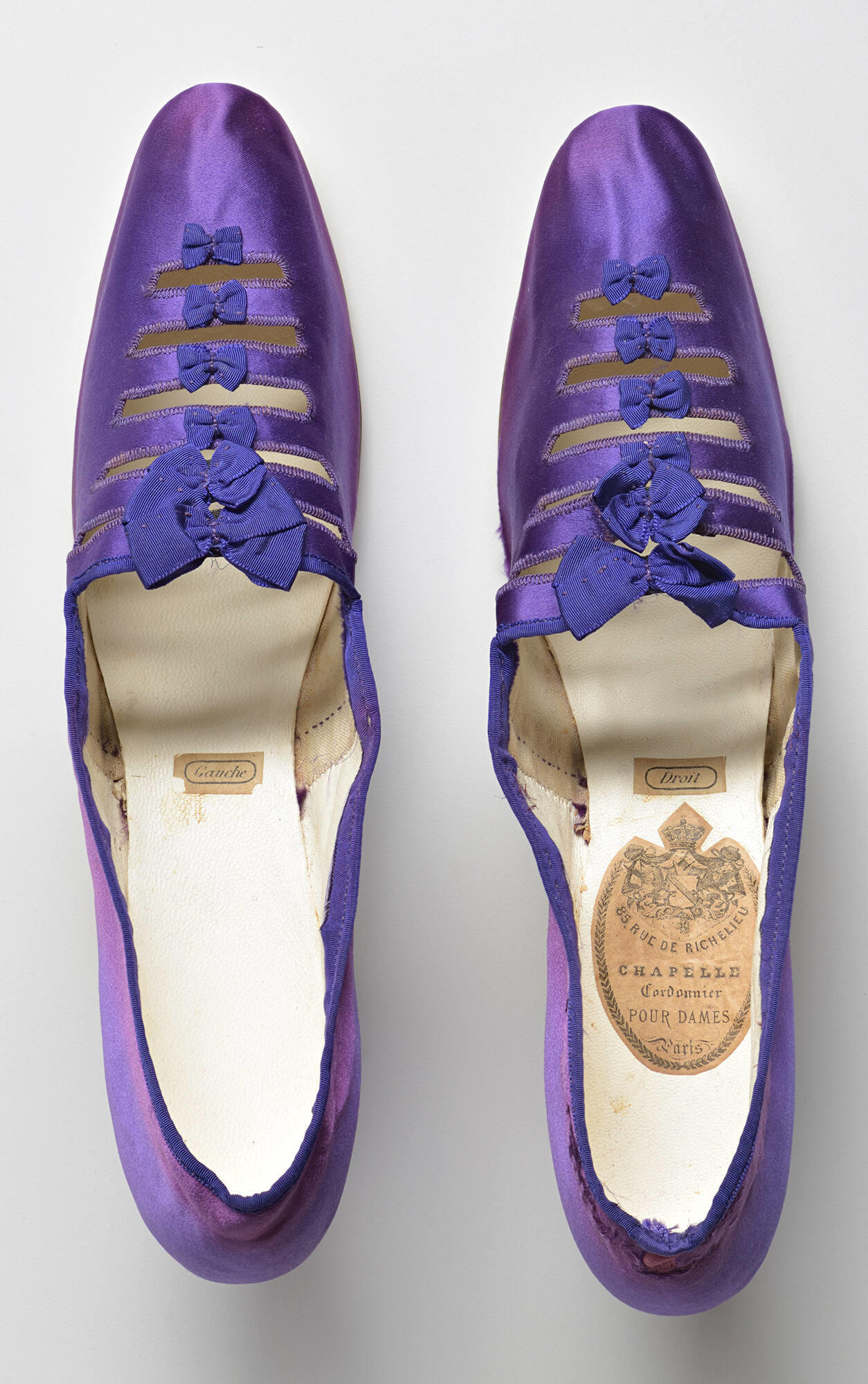 A pair of purple silk shoes seen from above with a label for gauche in the left shoe and droit in the right shoe. In the right shoe there is also a label for the shoemaker Chapelle, Cordonnier pour dames.