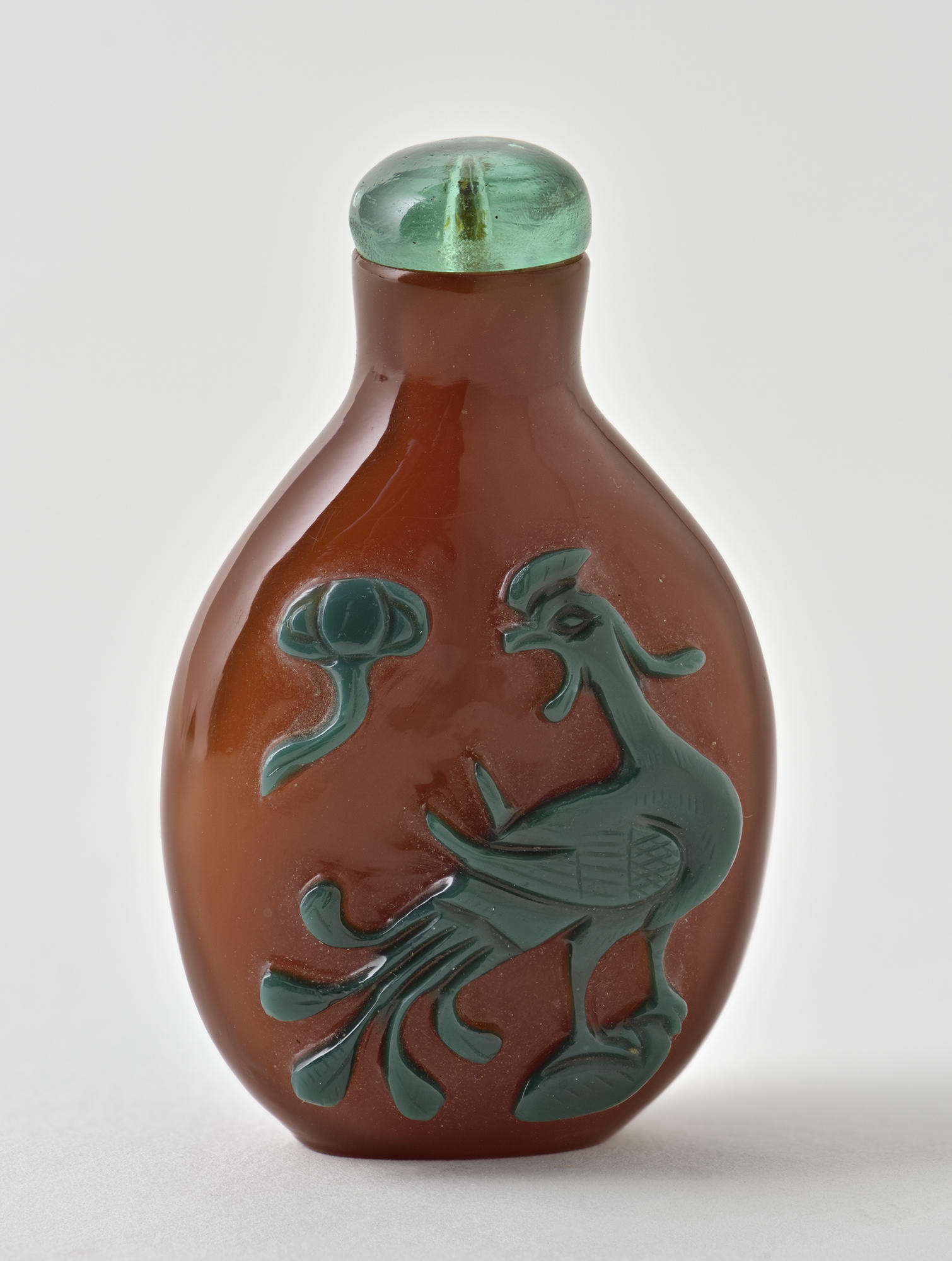  A red glass oval-shaped snuff bottle with a blue-green glass topper. There is a phoenix and sun and cloud motif in dark green cut glass.