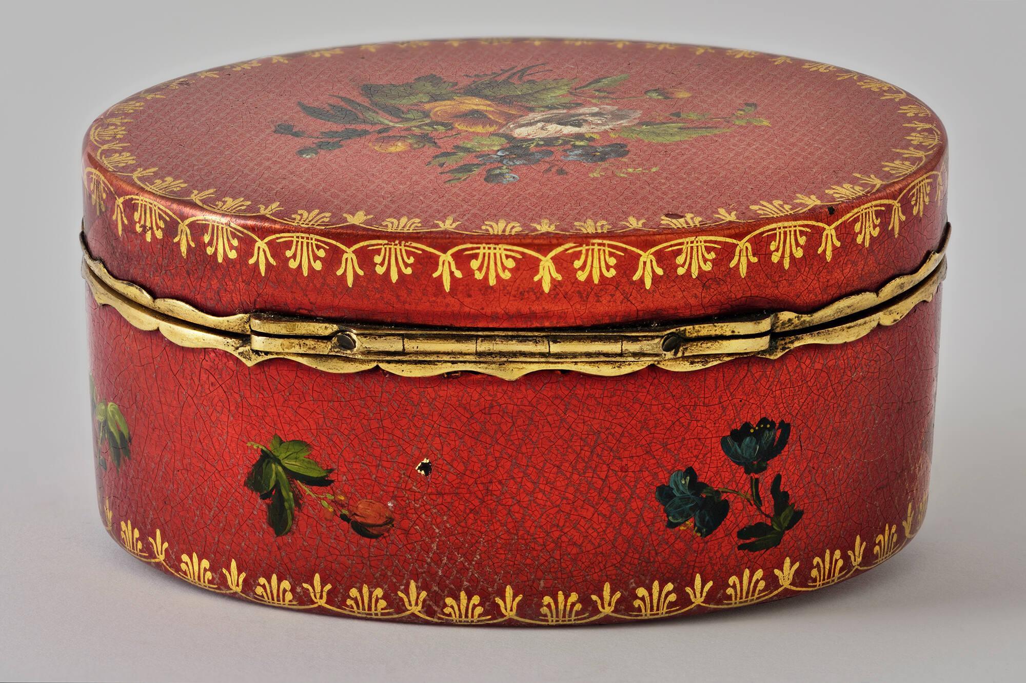 An oval red snuff box with a metal hinge and gold and floral decoration, in the Little Salon of the Gardner Museum.
