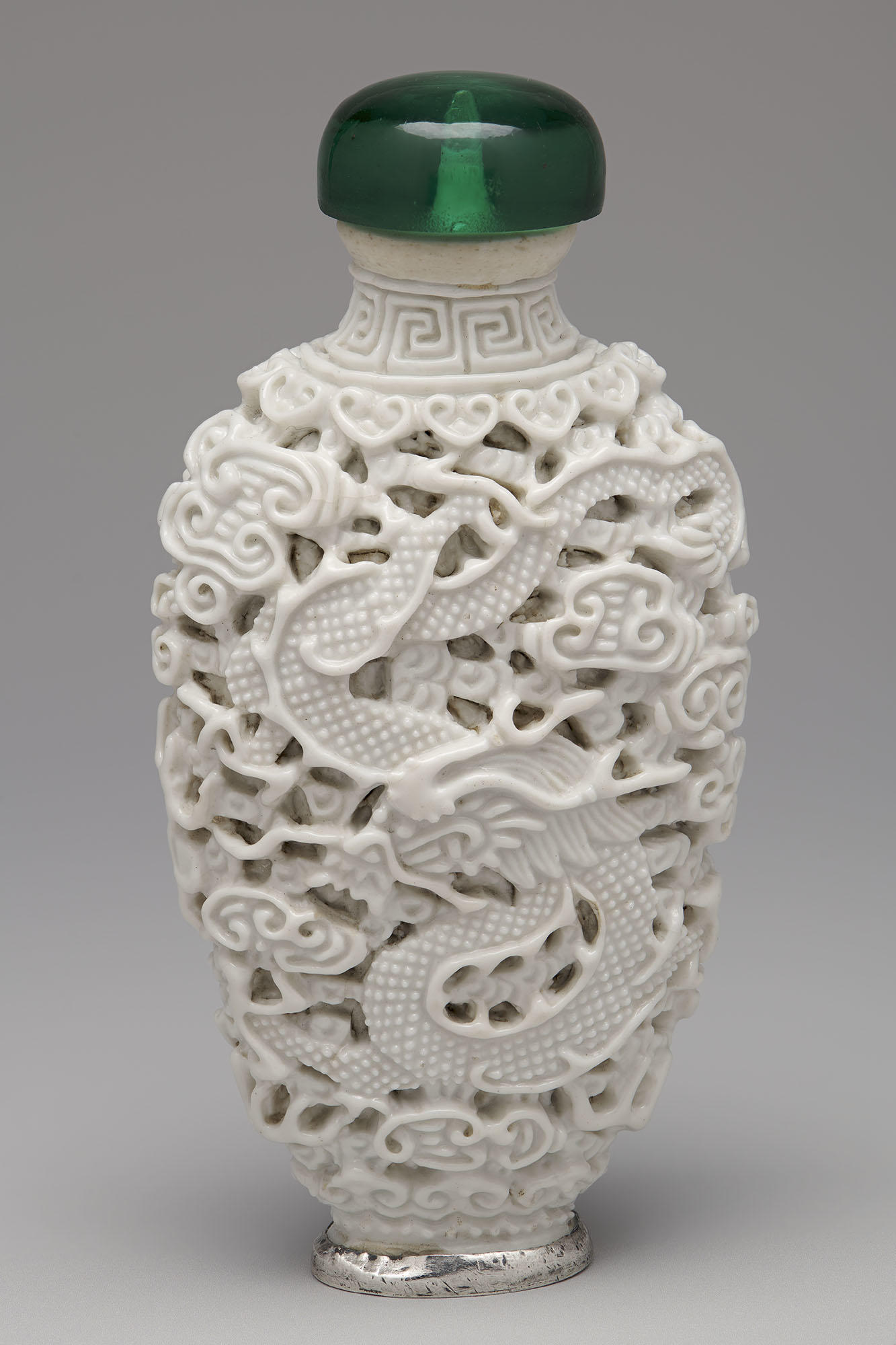 An oval-shaped white snuff bottle with a green stopper and silver rimmed foot. Carved into its surface is a dragon among clouds. 