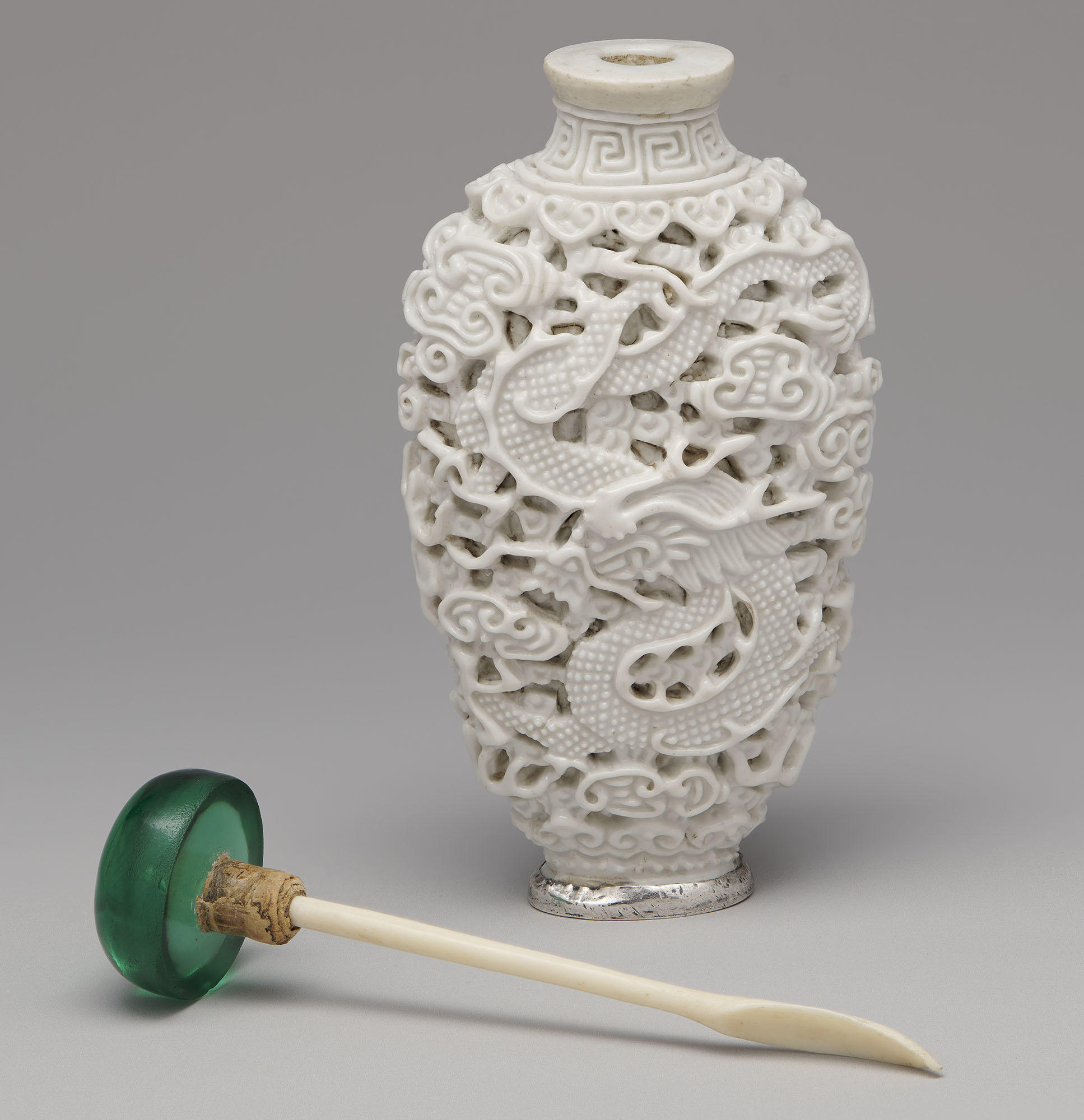 An oval-shaped white snuff bottle, with a silver rimmed foot, with the green stopper laying next to the bottle. The green stopper has a bone spoon attached to it. Carved into the surface of the bottle is a dragon among clouds. 