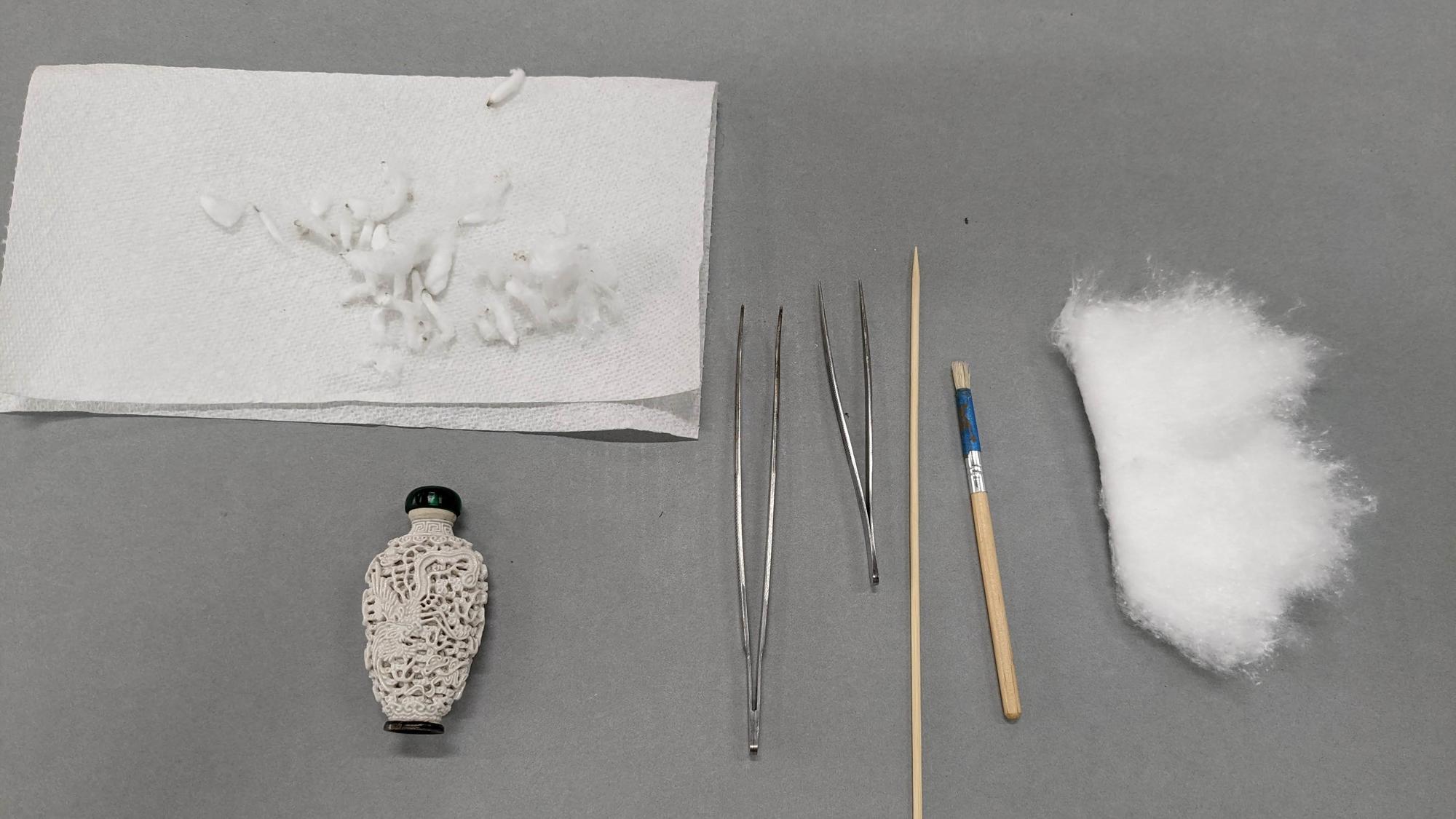 A white snuff bottle with a green lid lies next to implements (from left to right: two sizes of tweezers, a bamboo skewer, a stiff short haired brush and cotton) used to clean the bottle’s reticulation. Above the bottle are some cotton swabs used to clean the dirt and grime from the reticulation. 