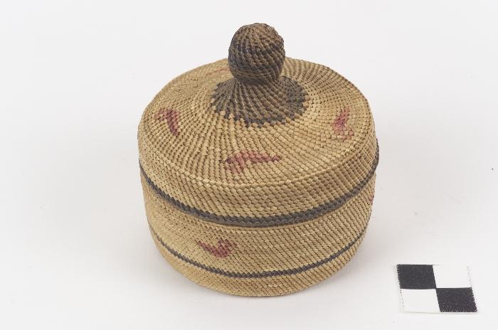 A cylindrical woven basket with a lid with a knob, decorated with a bird motif, made by a Makah weaver.