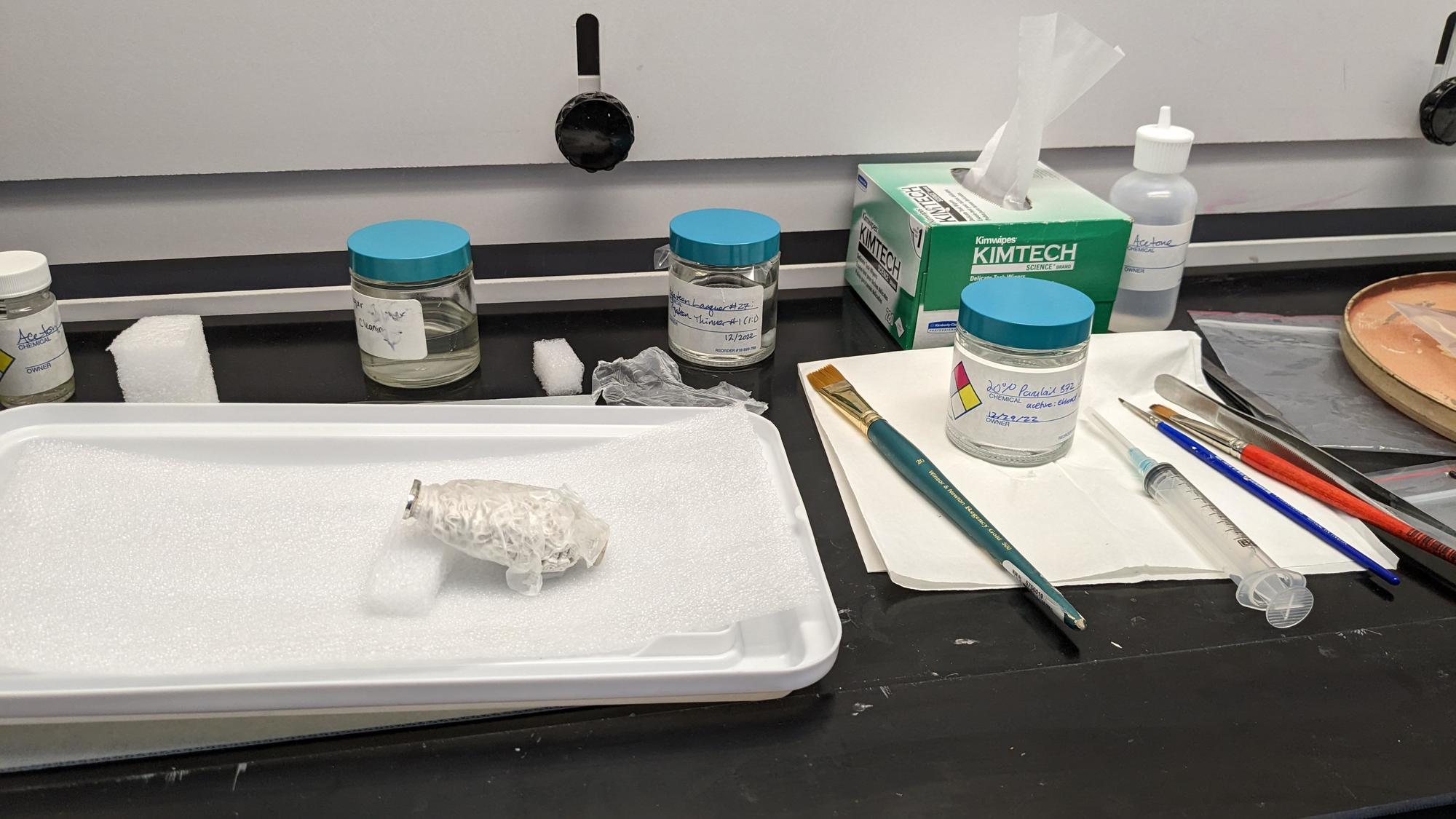 A snuff bottle is propped up on foam in a fume hood, with the silver foot exposed. To the right of the bottle is a solution of 20% Paraloid B-72, used to stabilize the cracked fragment of the foot, a syringe, brushes and tweezers. 