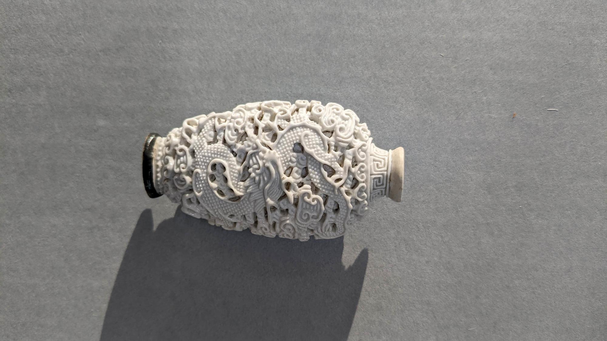 An oval-shaped white porcelain snuff bottle. Dramatic lighting from the left side of the bottle, highlights the deep and intricate carving of the dragon and clouds in the porcelain. 