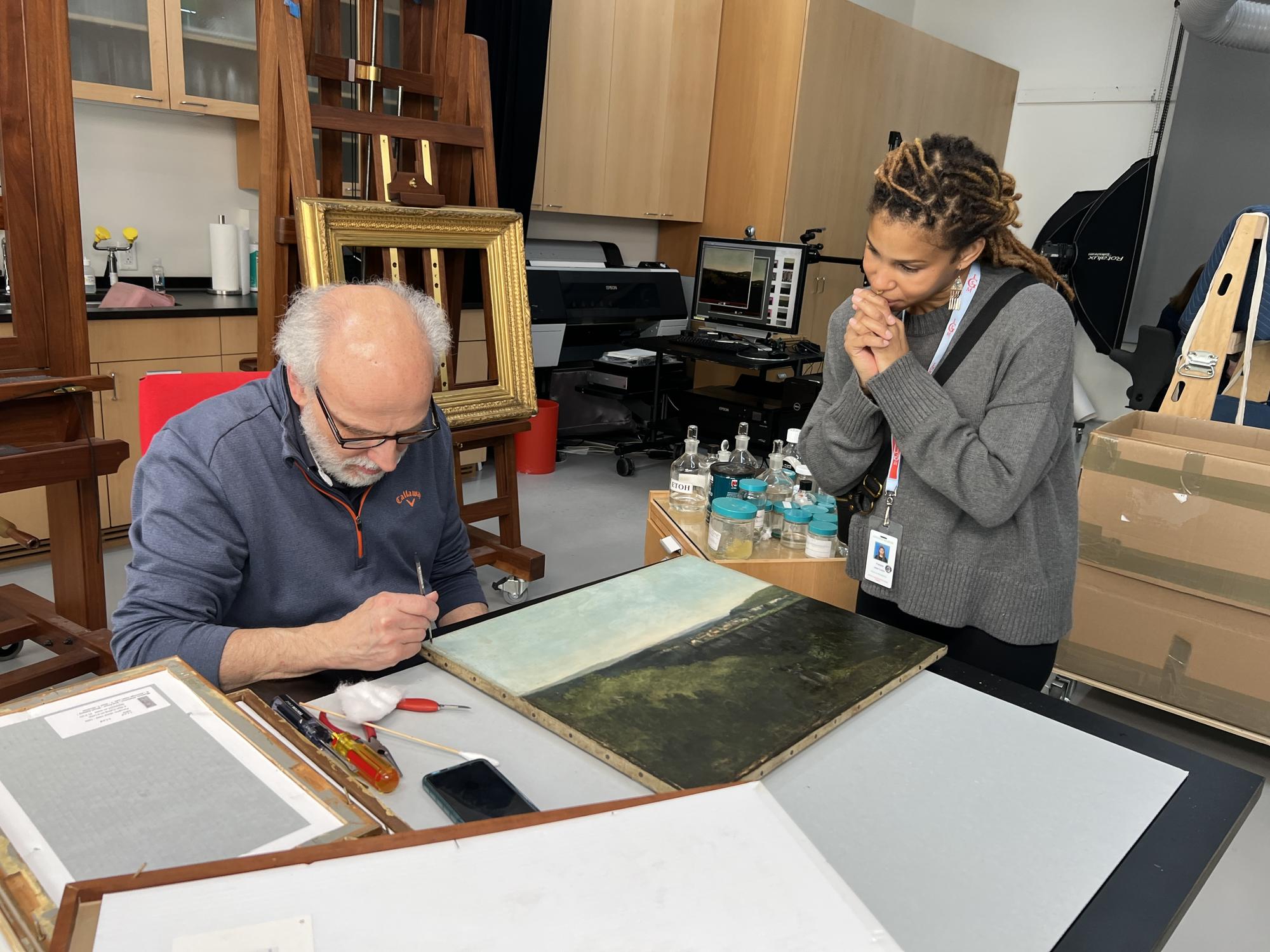 Conservator Gianfranco Pocobene removes varnish from A View Across the River by Gustave Courbet while Fabiola Jean-Louis looks on, 2022.