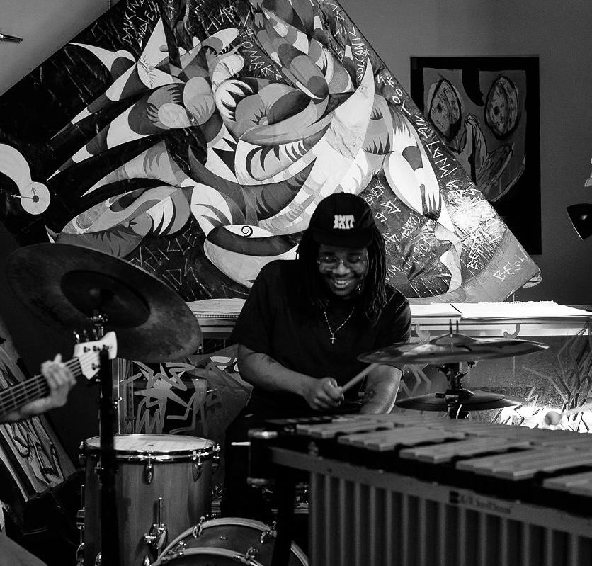 Black and white image of Maliq Wynn Johnson sitting in front of a large art piece. 