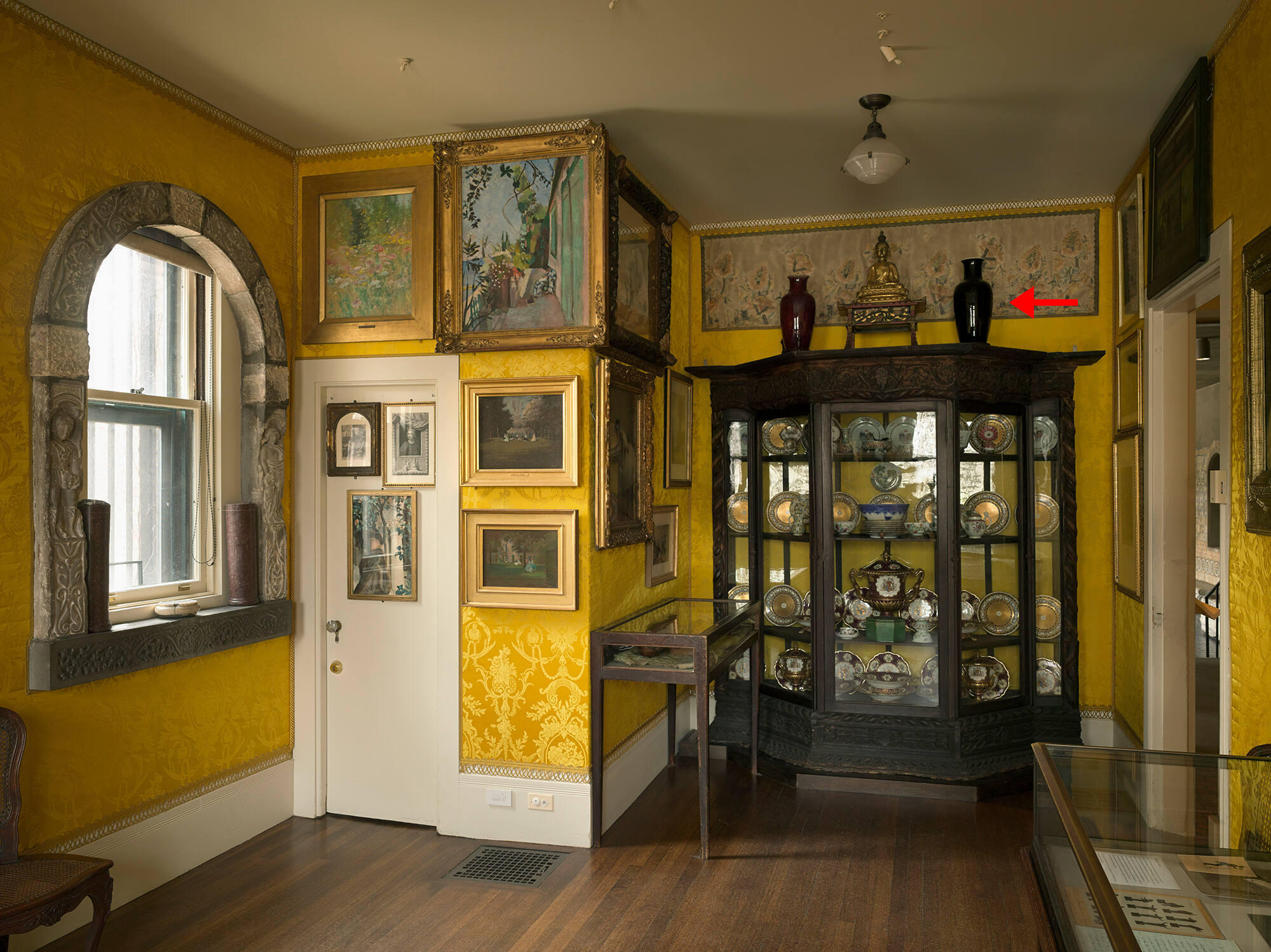 A room with yellow upholstered walls and a tall wooden cabinet with glass doors. A red arrow points to a black vase on top of the cabinet