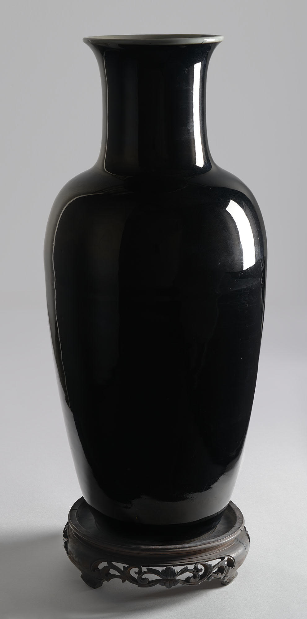 Full view of black vase, taller than it is wide, with a rim that turns outward, a slim neck, and a shoulder that slopes gradually to a narrower base on a wooden stand.
