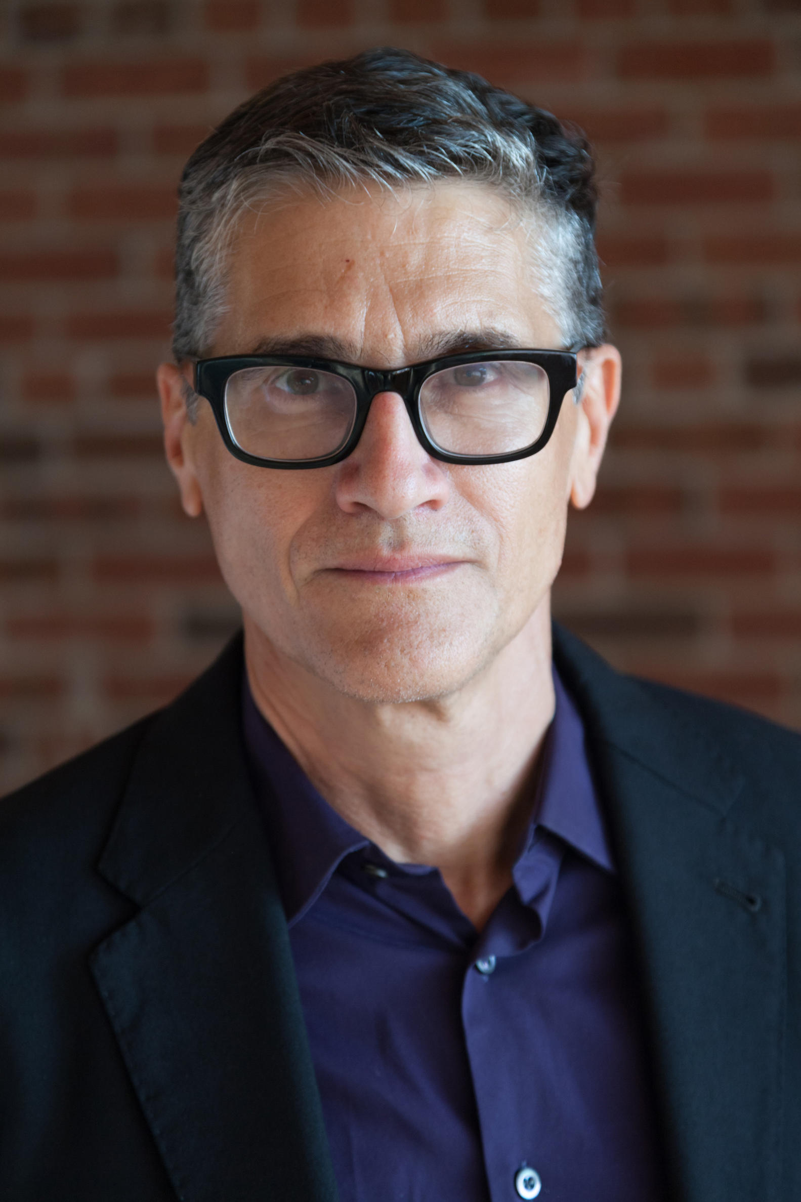 Headshot of man with greying dark hair and glasses wearing a purple shir and black jacket 