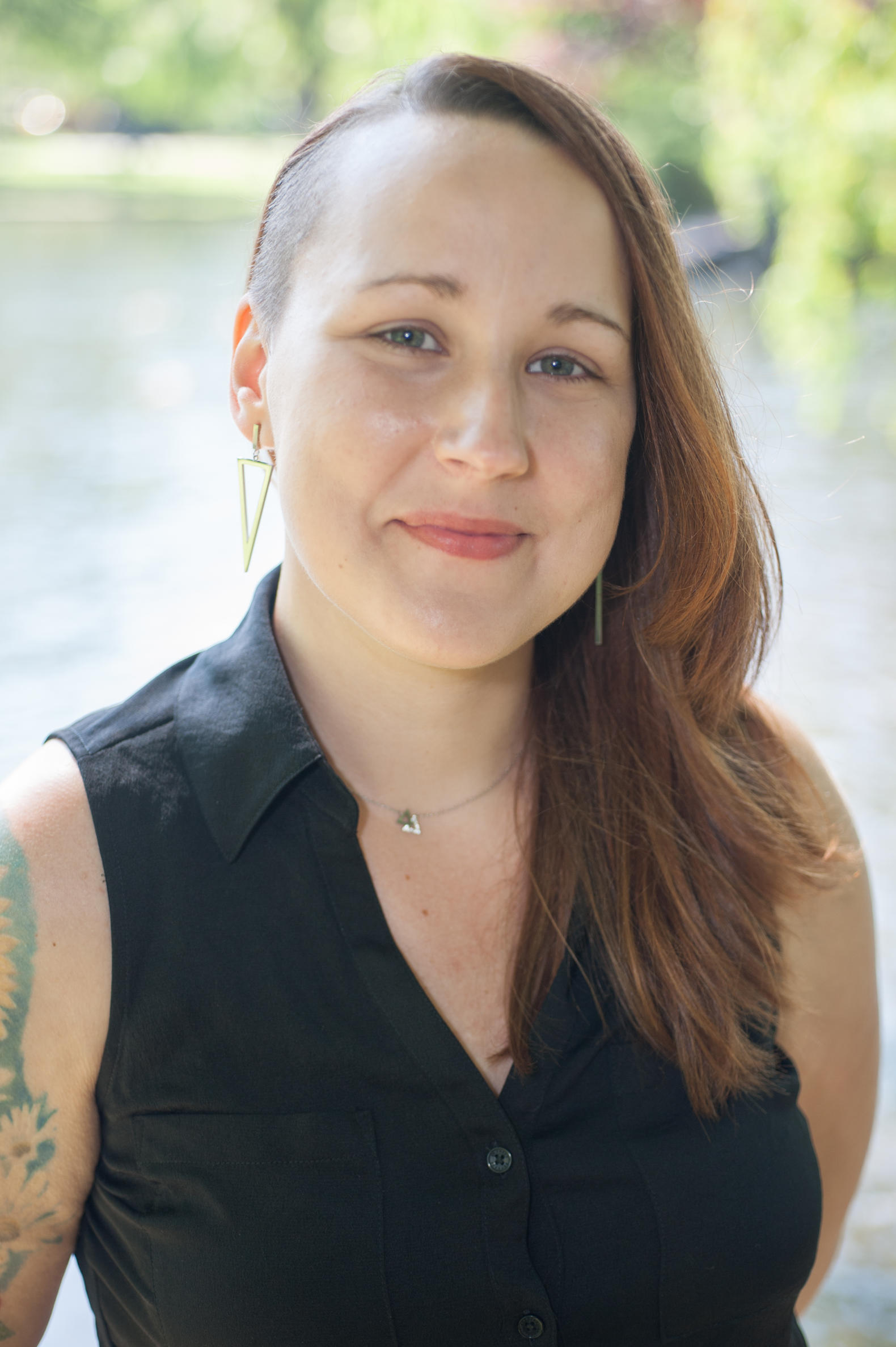 Woman with partially shaved head and long hair, wearing a sleeveless black top with tattoos on her right arm. 