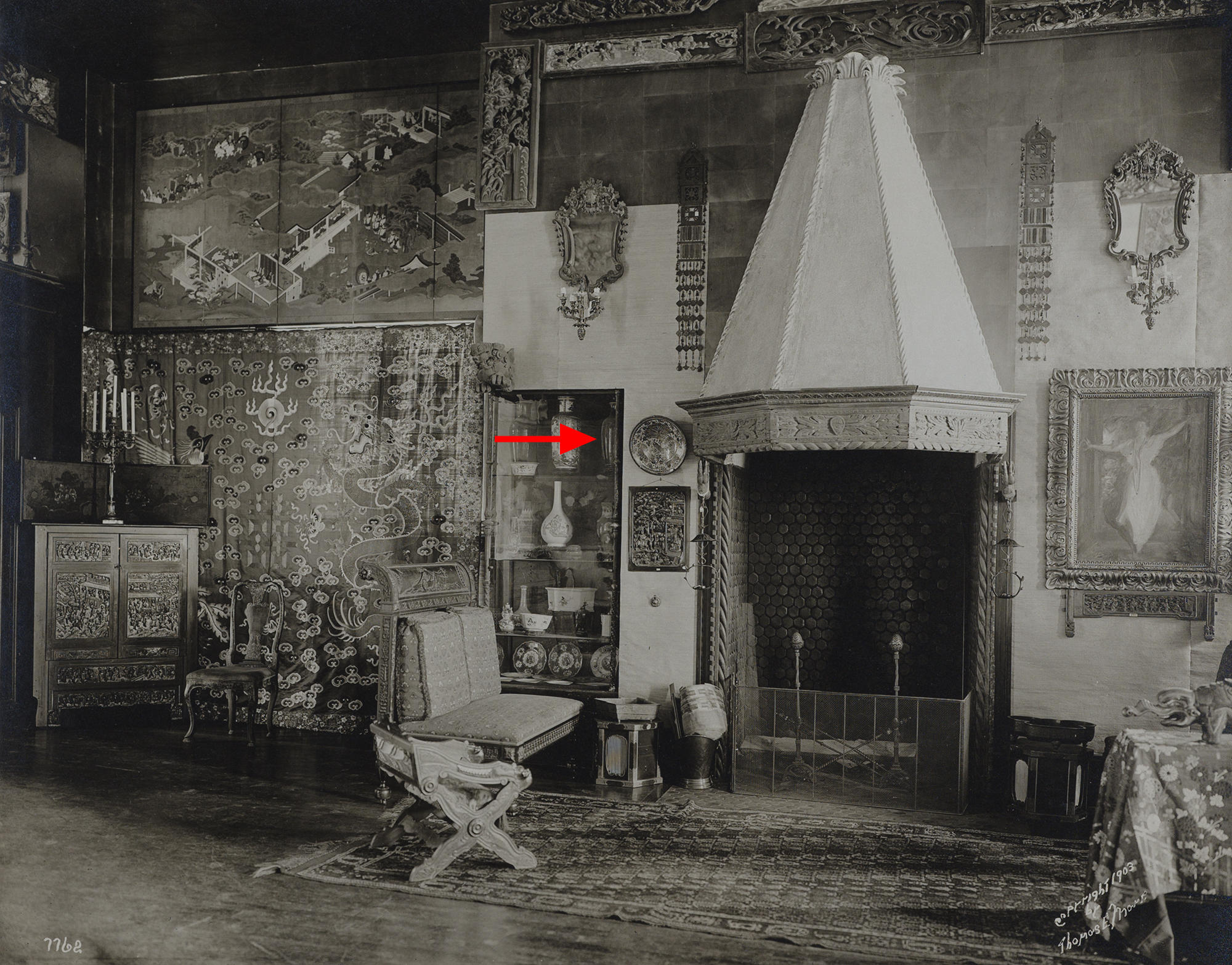 Black and white photograph of a gallery wall with Japanese screens and a Chinese textile. A red arrow points to a vase on the top shelf of a glass cabinet. 