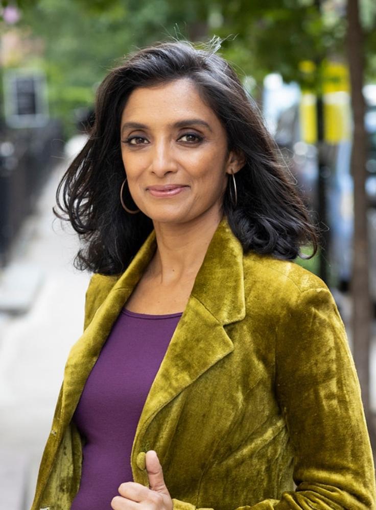 Woman with shoulder-length brown hair with a purple top, green jacket standing in front of trees outdoors. 