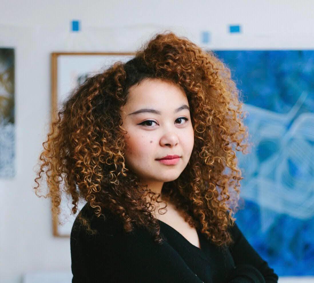 Asian woman with curly redish-black hair in a black top, standing in front of a blue piece of artwork, looking at the camera