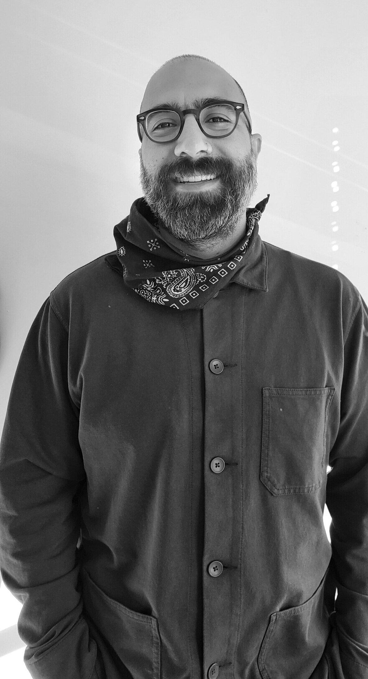 Black and white photo of a man with dark glasses, beard and a bald head with a dark shirt and bandana tied around his neck. 