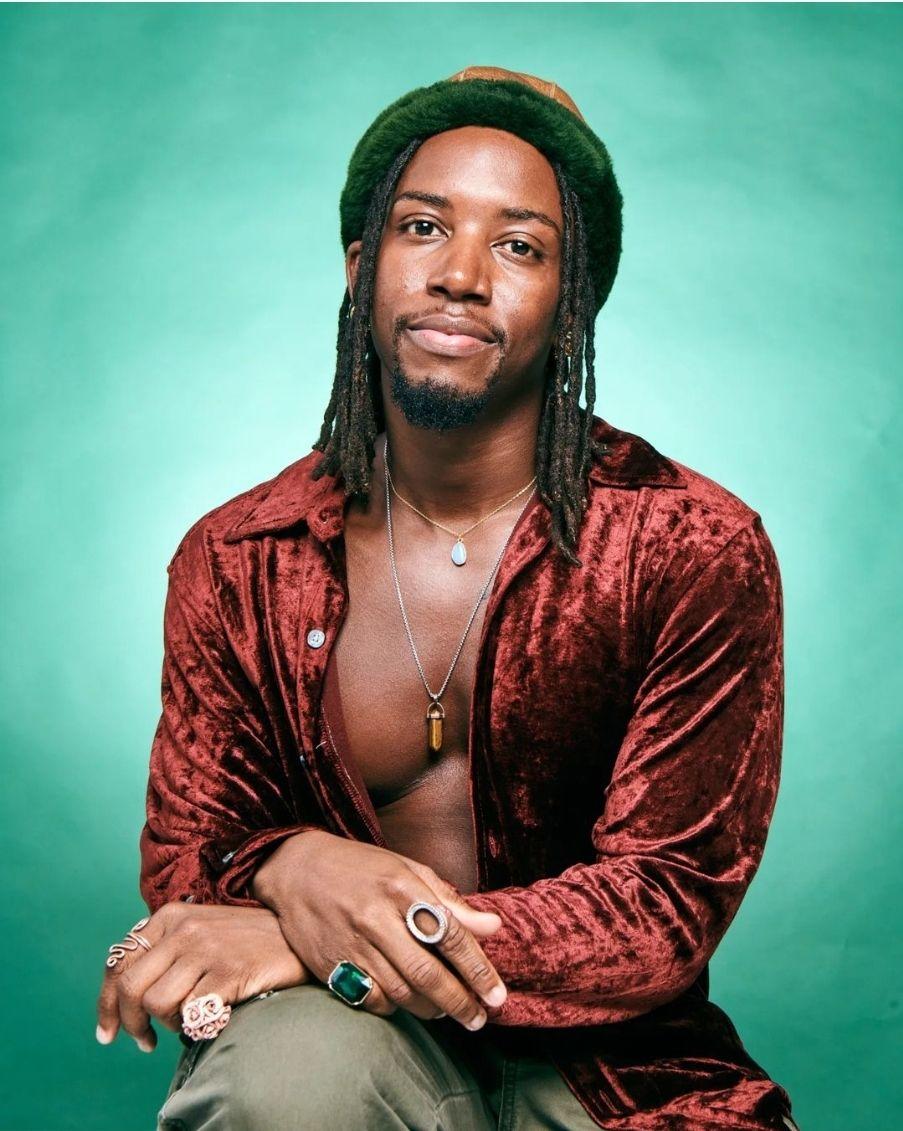 African American person with dreadlocks, hat, facial hair with a velvet top in front of a green backdrop