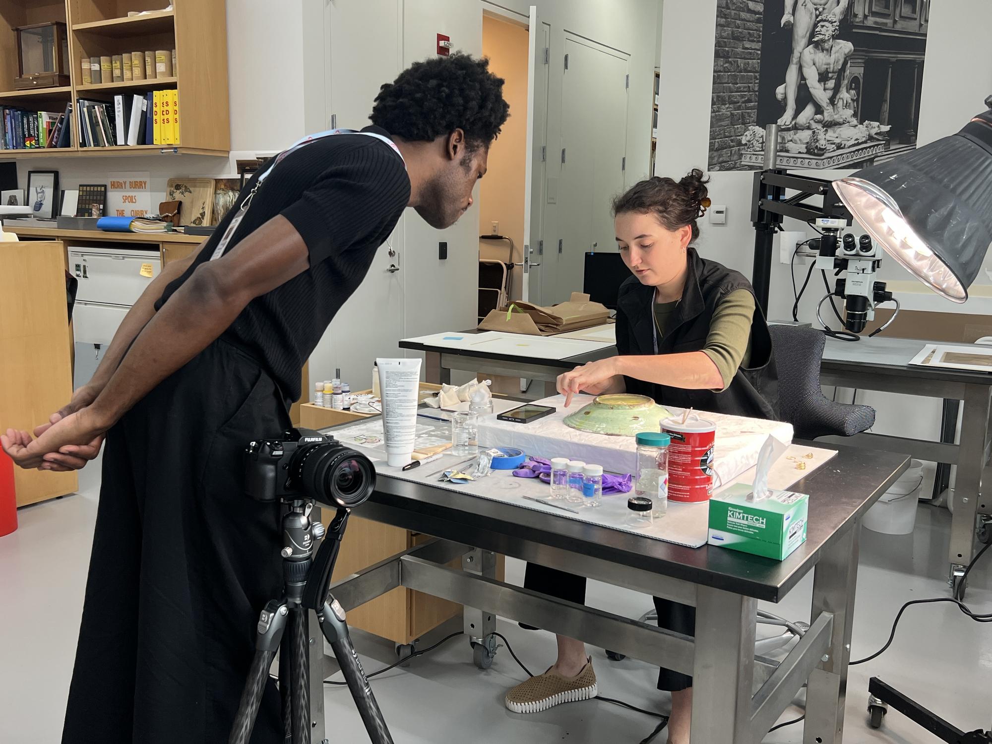 Man in black shirt looking at art on a desk with a woman in the conservation department 