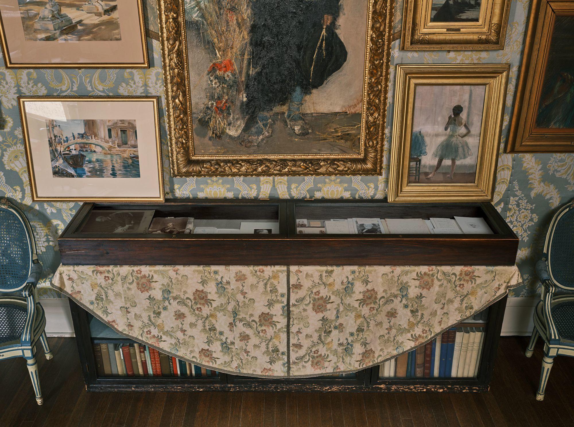 A glass covered case with photographs, papers, and books in a room with paintings on the wall.