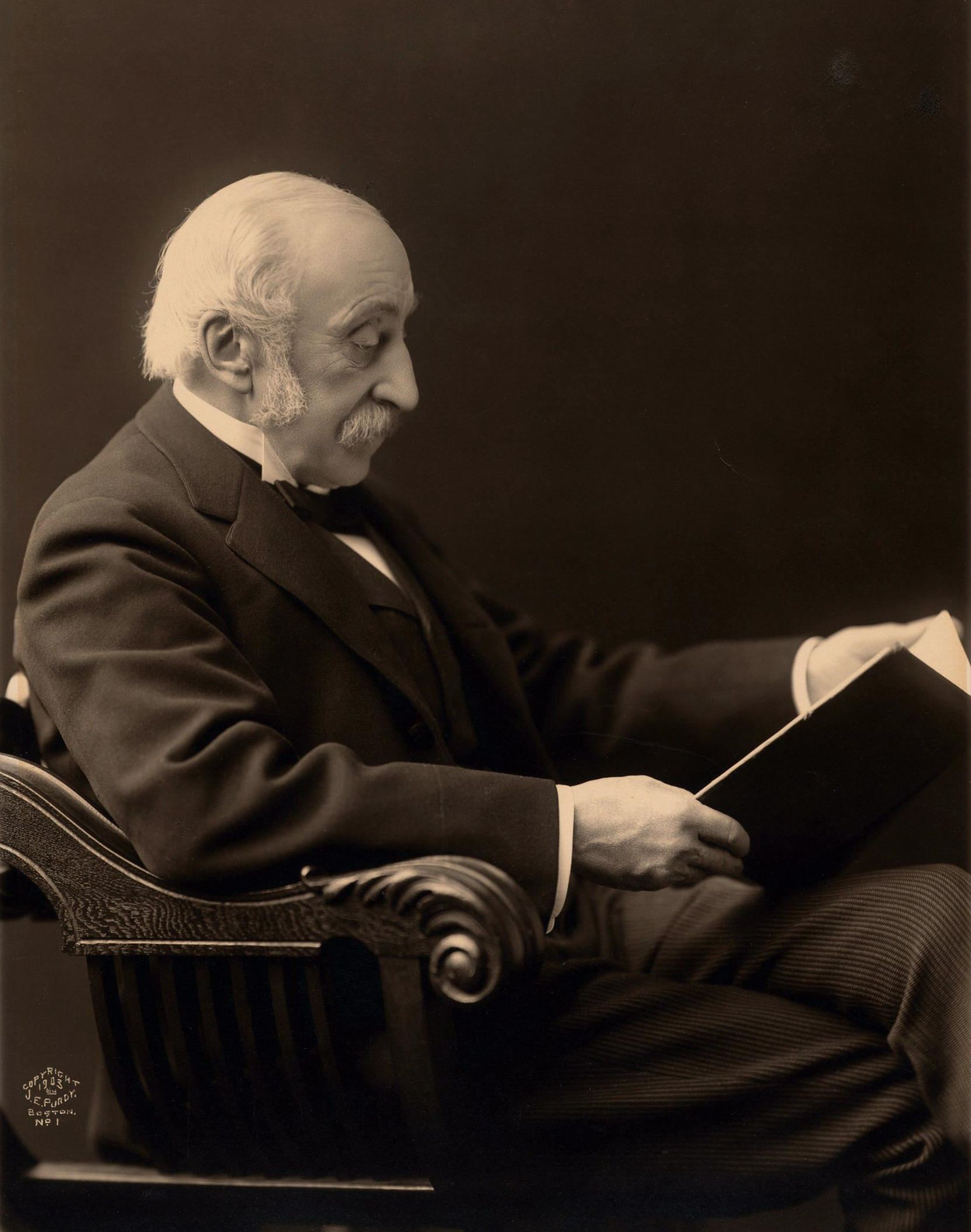 A black and white photograph of a seated white man with balding white hair in profile reading a book.