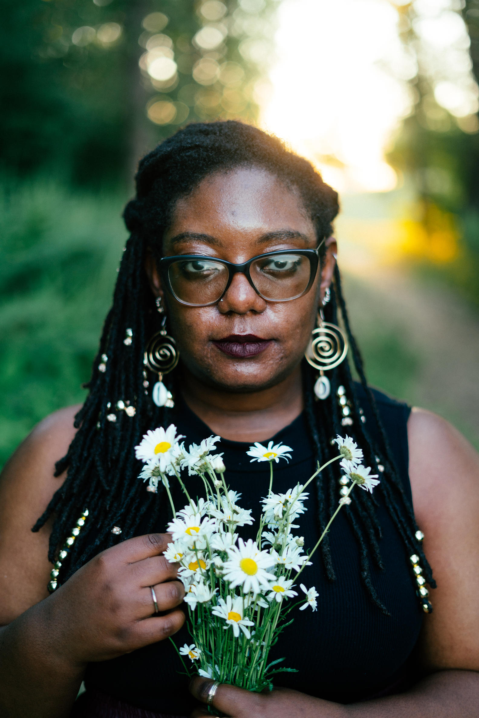 Afro, Carribean, Latina woman with long dread-locks and glasses holding a a bunch of daisies. 
