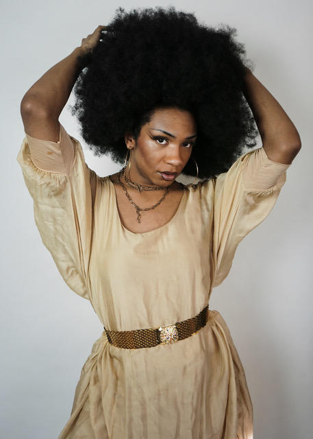 Person with an afro, tan dress with a belt, looking forward