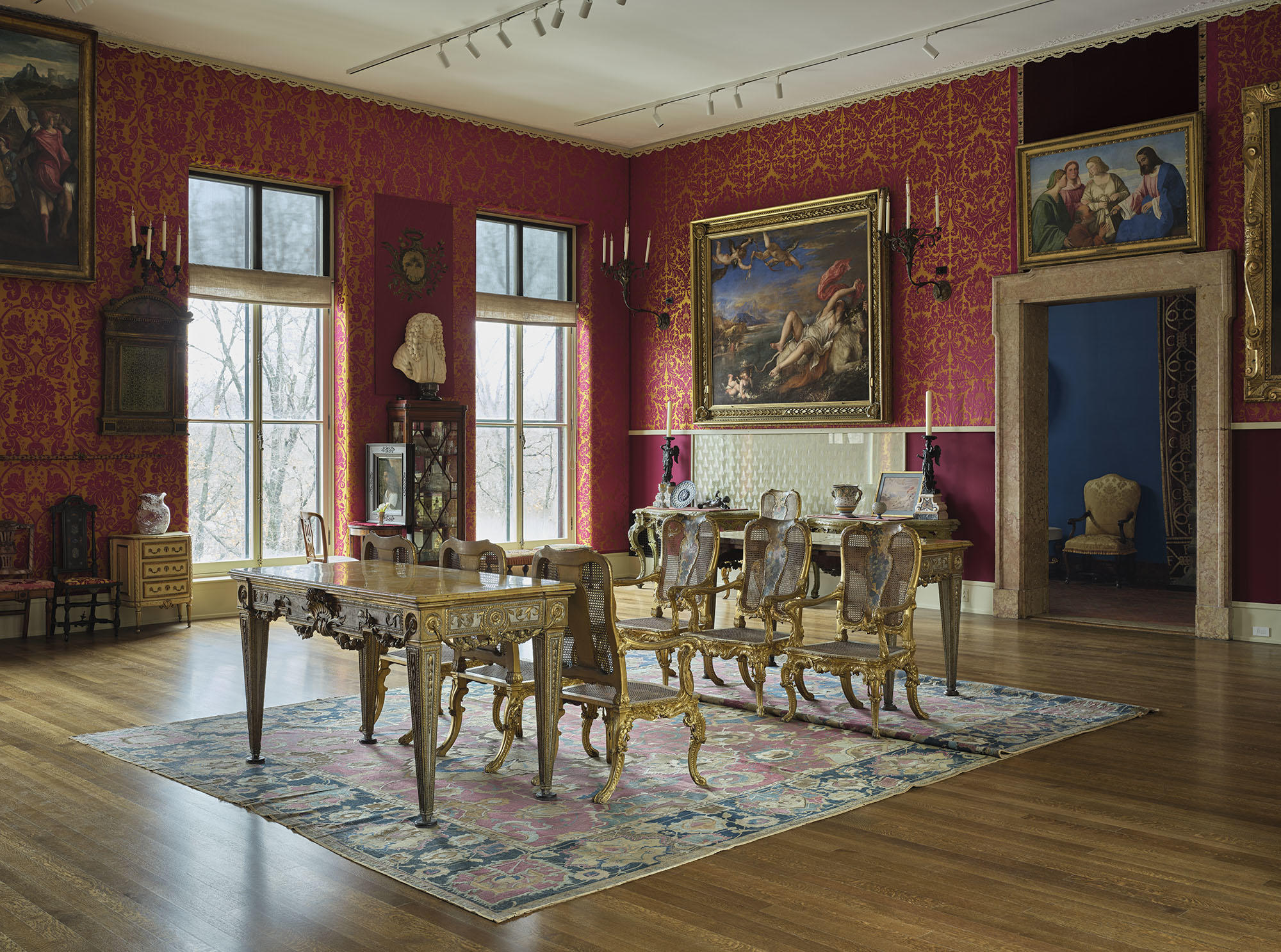 A color photograph of the Gardner Museum’s Titian Room showing six of the giltwood chairs displayed between two tables on top of a large Persian rug, with a number of large frames hung in the background on the red and yellow floral walls.
