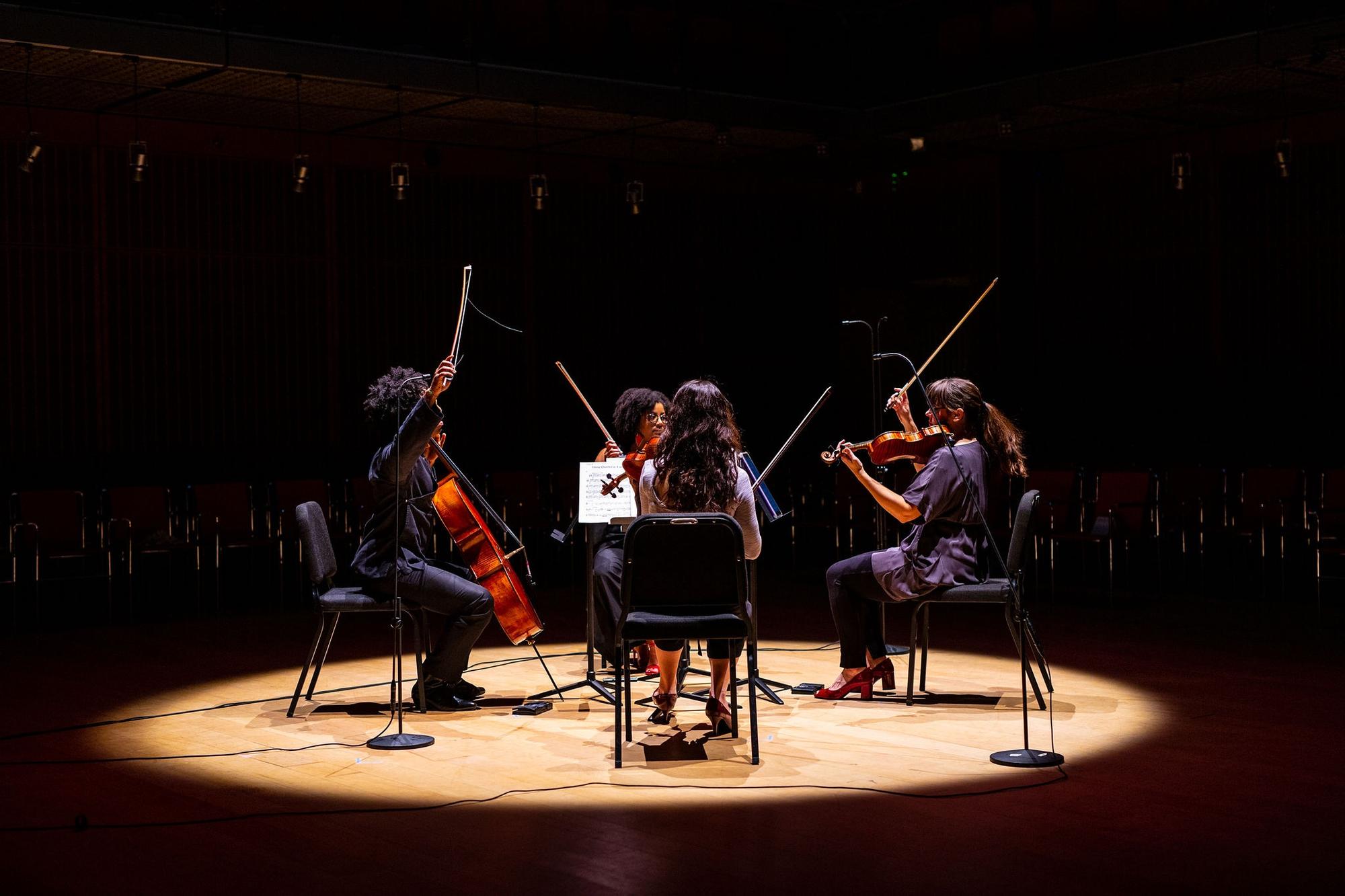 Four femal muscians sitting on chairs within a circle of light, facing one another playing string instruments. 