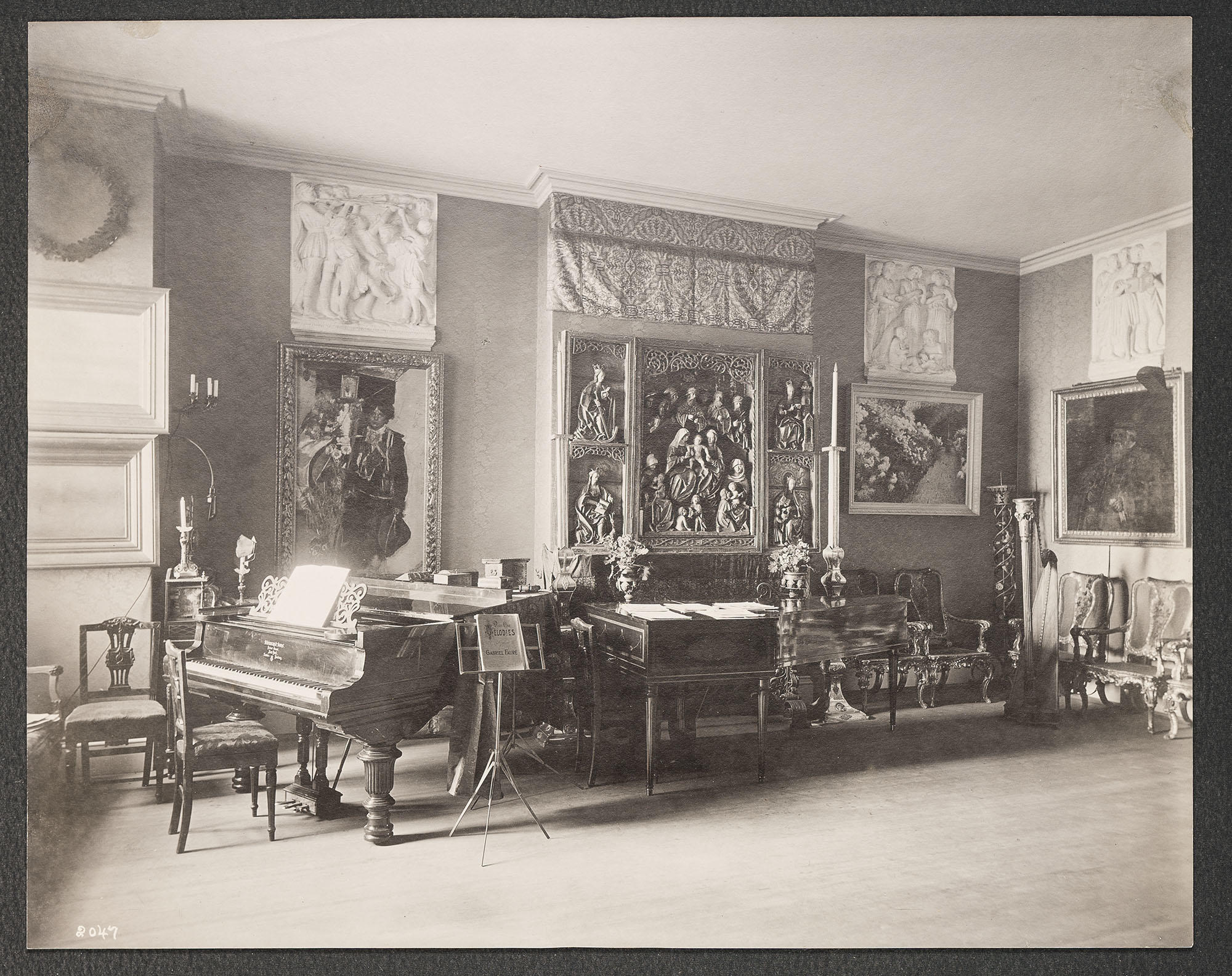 A black and white photograph of Isabella’s Music Room at 152 Beacon Street, Boston, with a piano, a harp, frames and sculptures on the walls, and the armchairs arranged around the back right corner.