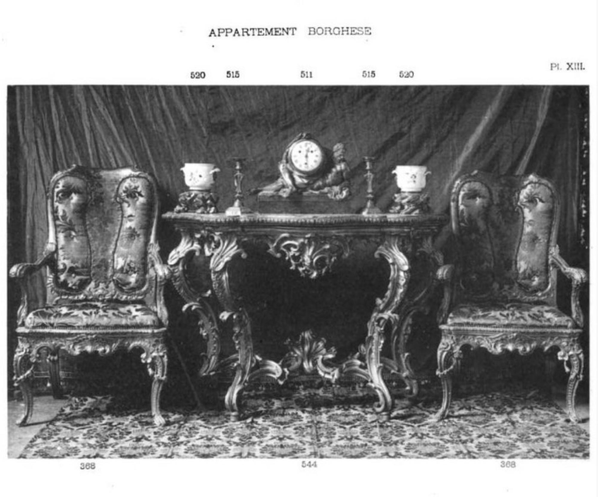 A black and white photograph from the auction catalog showing two of the armchairs placed on either side of a small table with a clock, candlesticks, and cups on it.