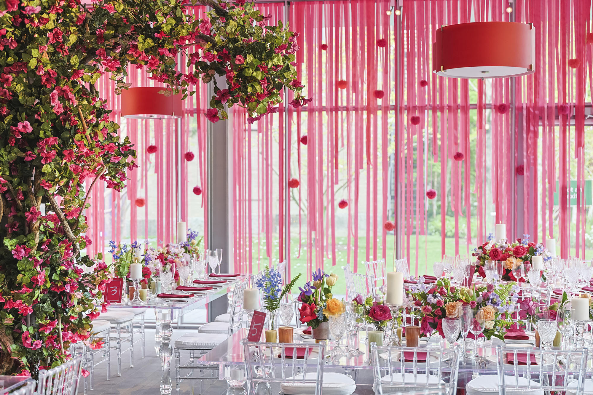 Room decorated with pink streamers falling from the ceiling with lush greenery and pink flowers surrounding white tables and chairs. 