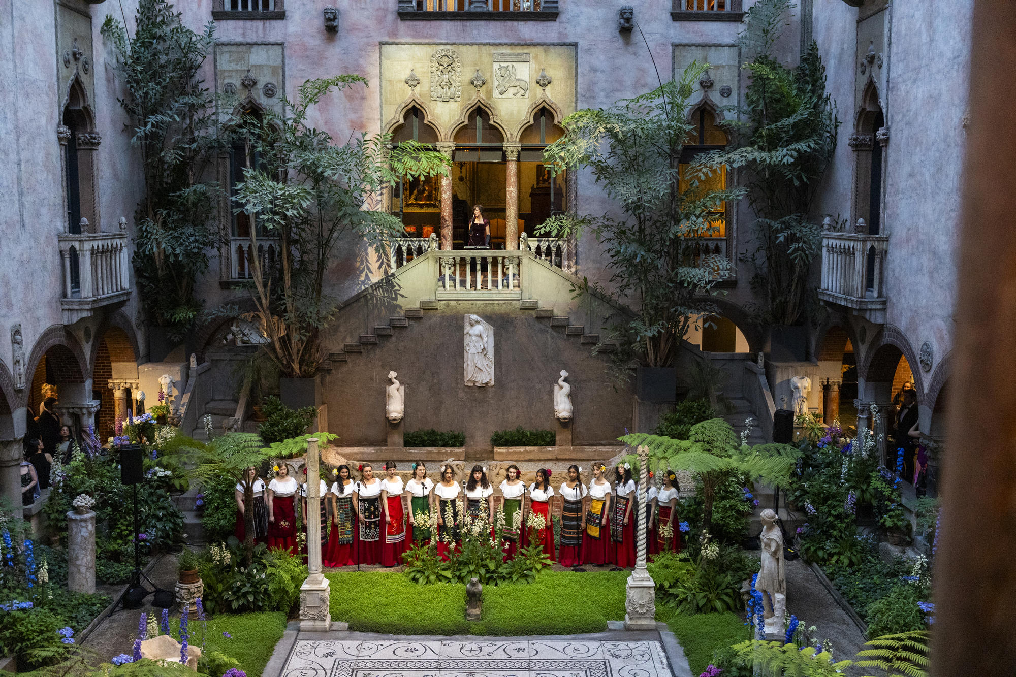 Color photograph of a group of singers on the ground level of the courtyard and a spot light on a woman standing in the balcony behind the singers. 