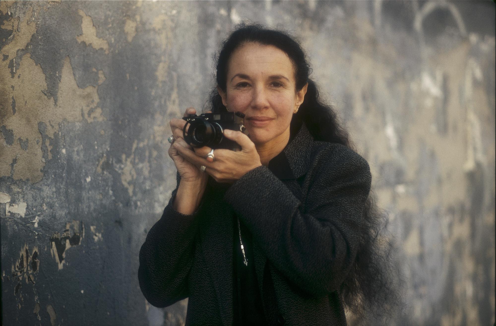 Woman with long dark wavy hair standing in front of a mixed colored wall holding a camera near her face looking confidently at the camers. 