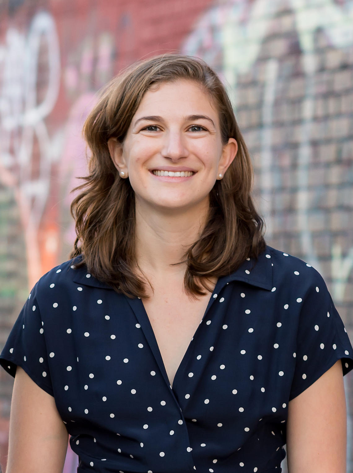 A woman with long, light brown hair weaing a navy blue polka dot top looking right at the camera smiling, standing in front of a brick, painted wall. 