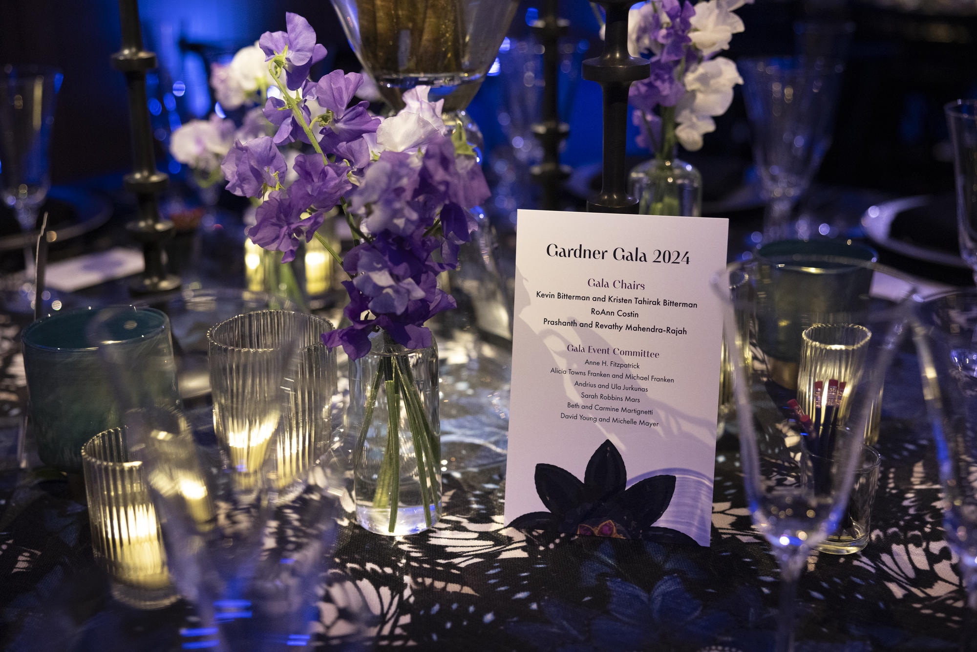 View of a placard with the names of the Gala Committee 2024 on a table with volitve candles, small vases of purple and white flowers on a black and white table cloth that resembles butterfly wings. 