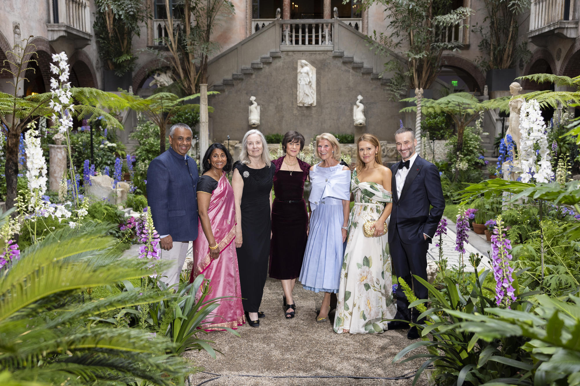 Seven elegantly attired people standing in the courtyard with lush plants around them. 