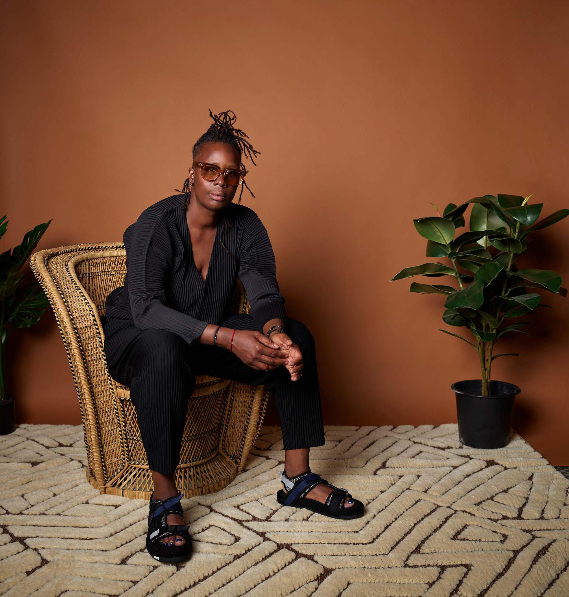 African American person with dread locks piled on top of her head in a black suit, sitting on a ratan chair, weaing glasses and sandles looking confidently at the camera. 