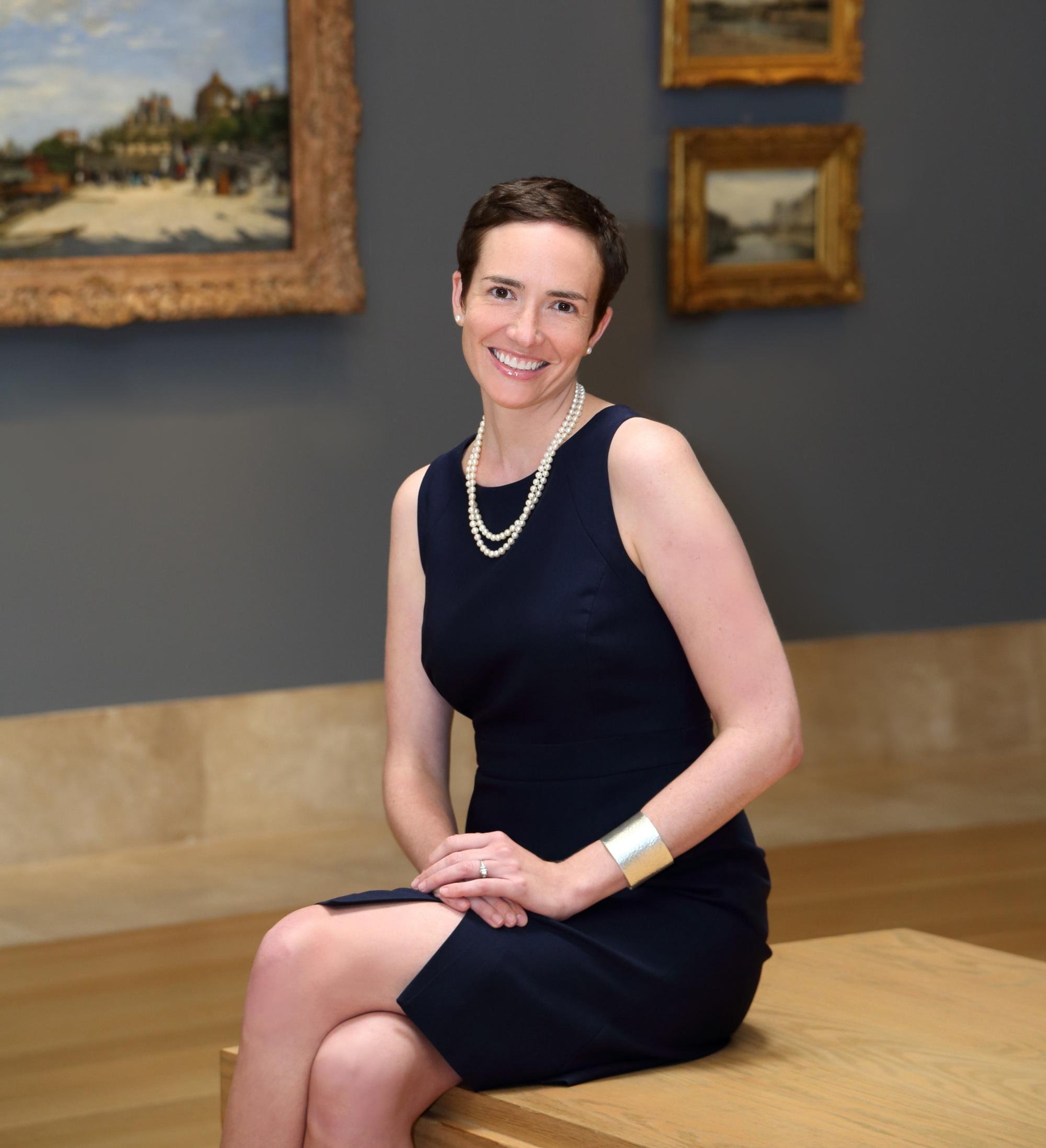 A woman with short brown hair wearing a sleeveless black dress and a sting of pearls sitting in front of paintings in a museum smiling at the camera. 