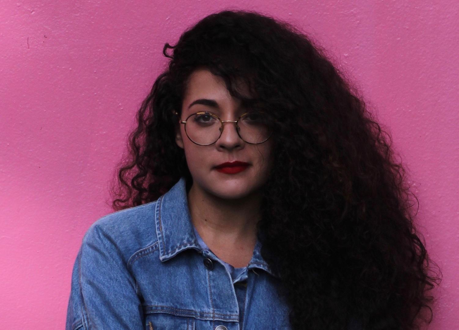 Woman with long, dark curly hair, glassed, red lipstick, jean shirt looking directly at the camera