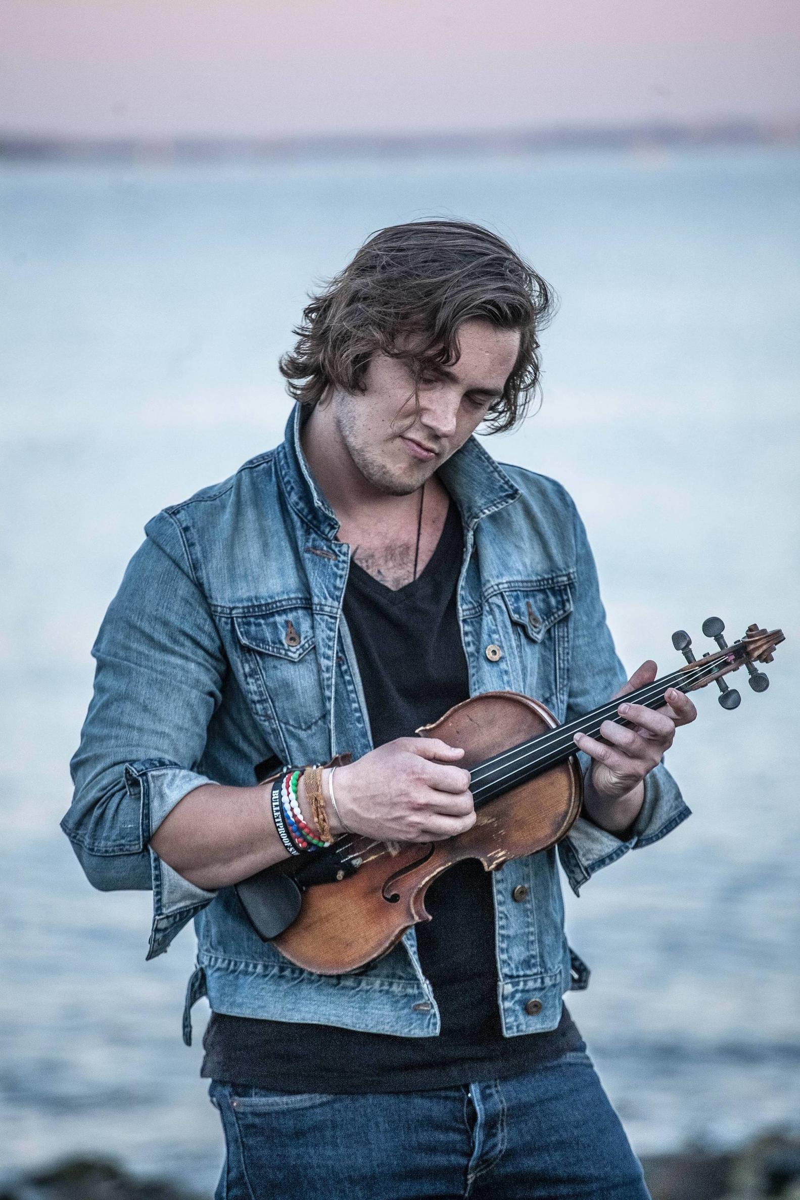 Young man in a black shirt and jean jacket holding a violin in front of a body of water. 