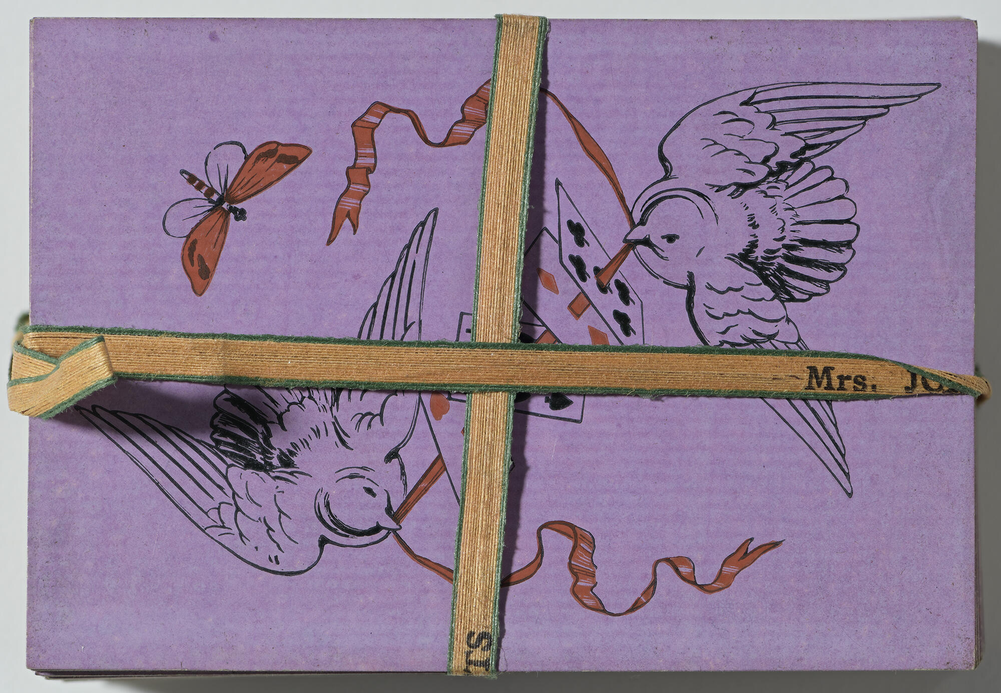An overhead view of a deck of playing cards, decorated with a design of two doves threading a red ribbon through four cards on a purple background, secured together by an orange ribbon that is personalized with Isabella’s name.