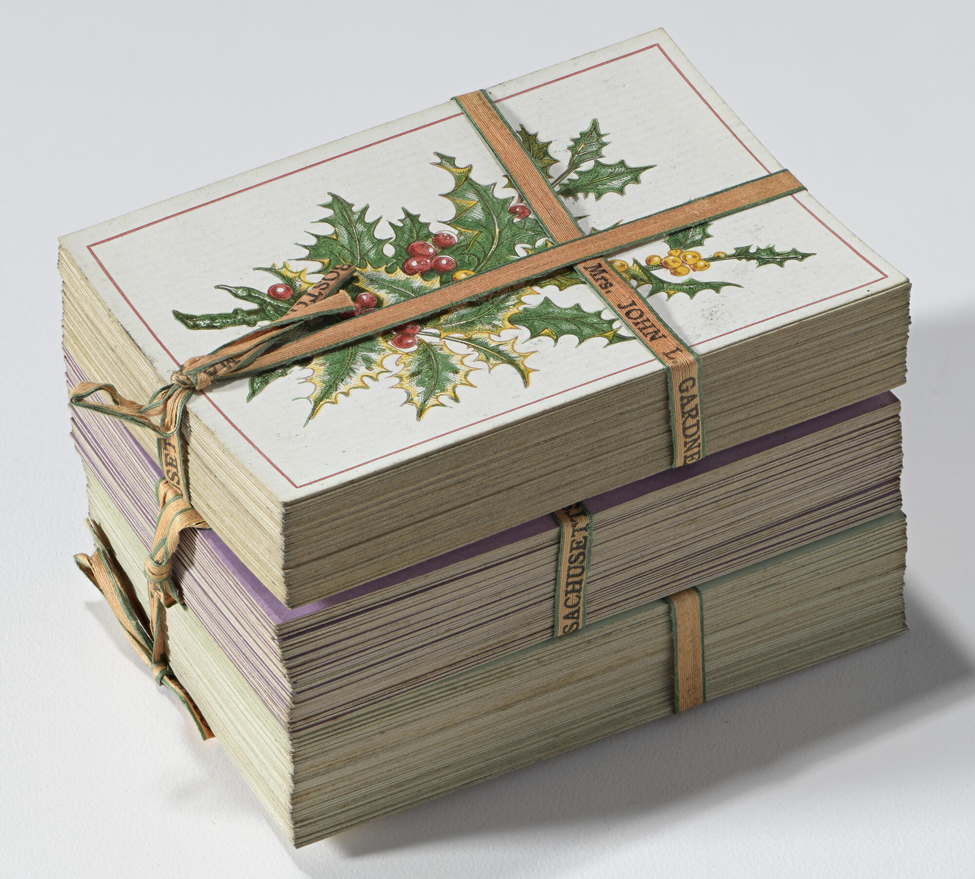 Three decks of playing cards, stacked on top of each other and secured with orange ribbon that is personalized with Isabella’s name, showing the back of the top deck which is decorated with a green, red, and yellow holly plant design.