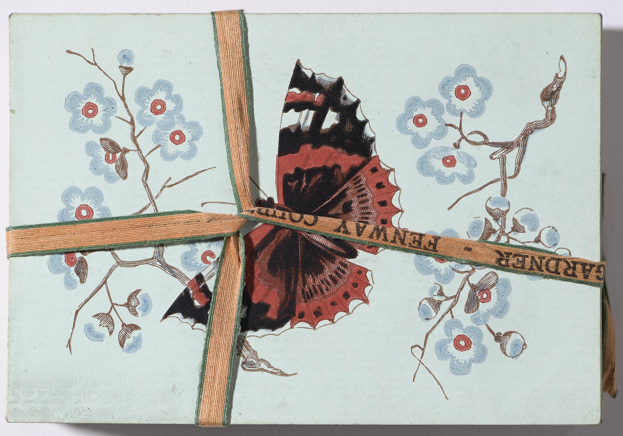 An overhead view of a deck of playing cards, decorated with a design of a red and black butterfly framed by blue and red flowers on brown branches on a light blue background, secured together by an orange ribbon that is personalized with Isabella’s name.