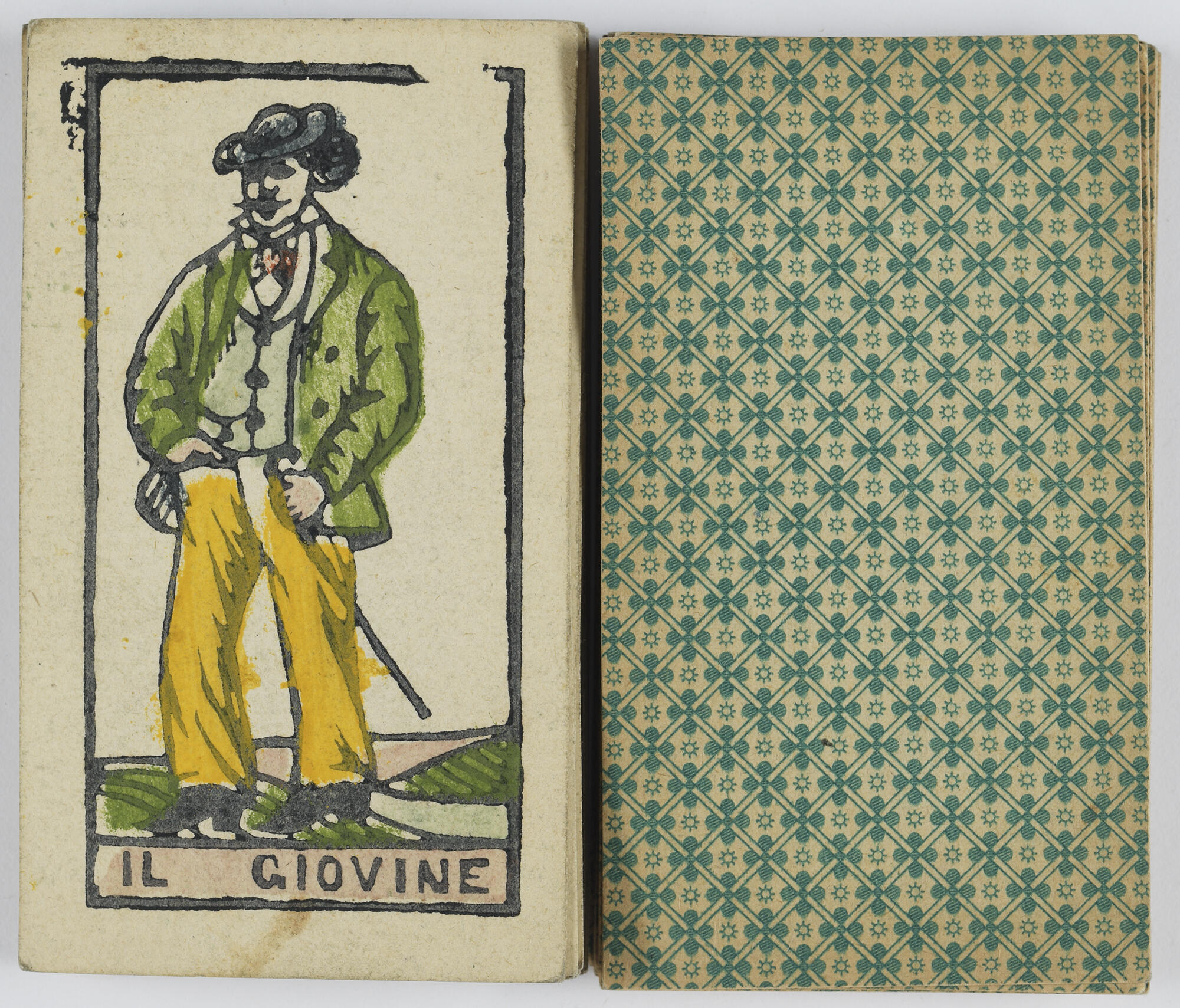  An overhead view of the ‘il giovine,’ or ‘young man’ card, showing a man in a black hat, green jacket, and yellow pants, with a mustache and the back of another card from the deck, decorated with a green pattern of quatrefoils, suns, and intersecting lines.