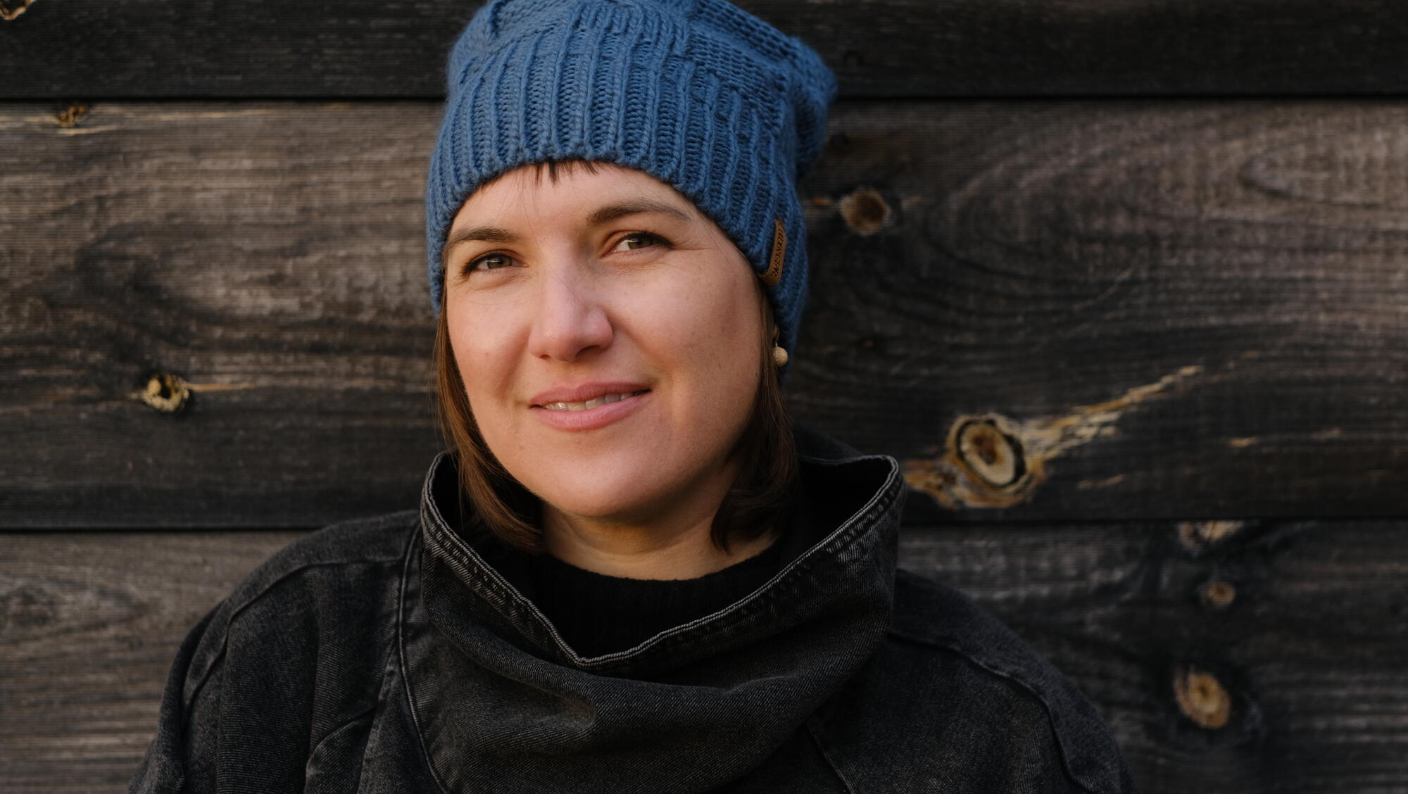 White woman with a blue knit hat in front of a wooden wall with a black top
