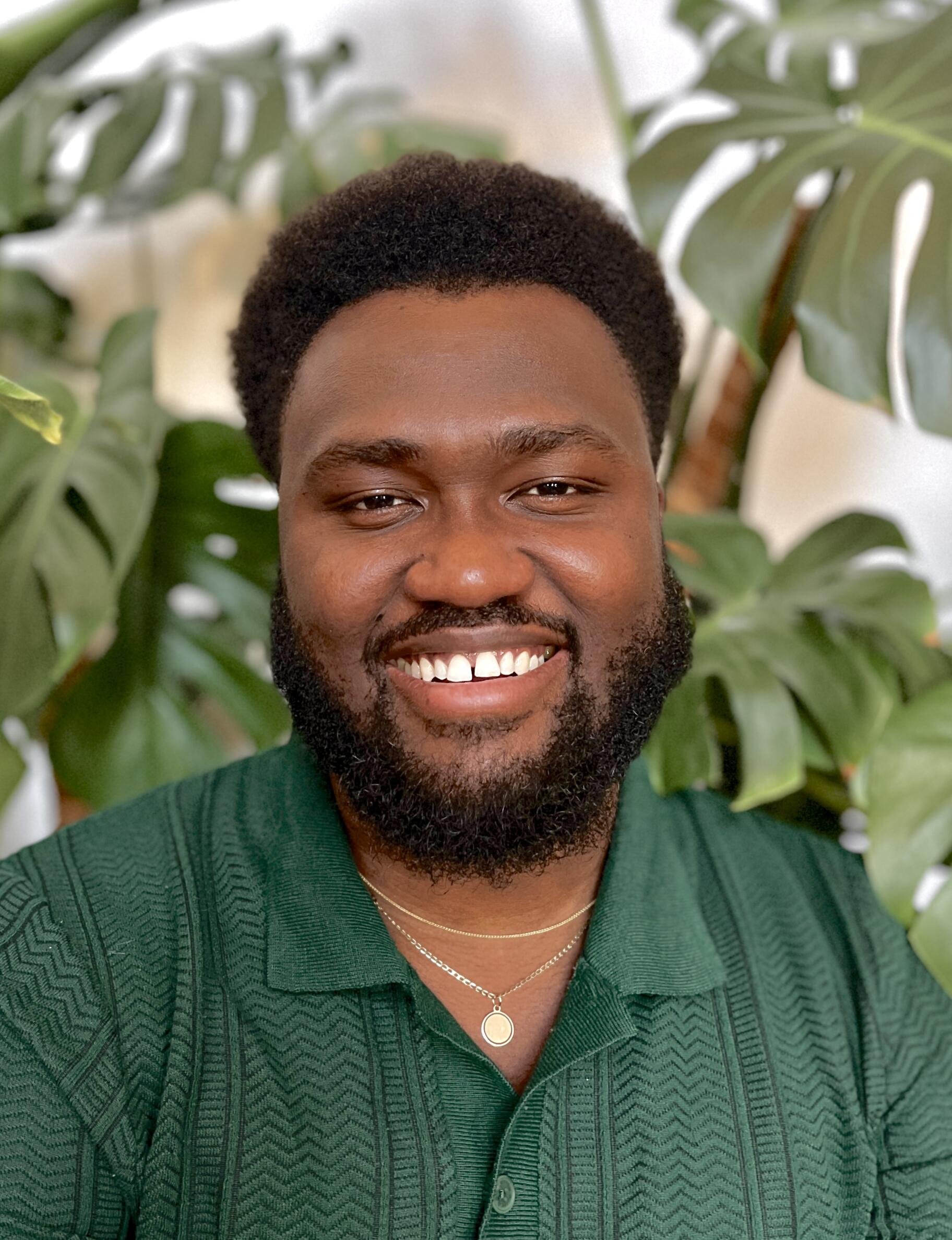 African American man with a big smile,looking directly at the camera, wearing a green shirt and sitting in front of a monstera plant. 