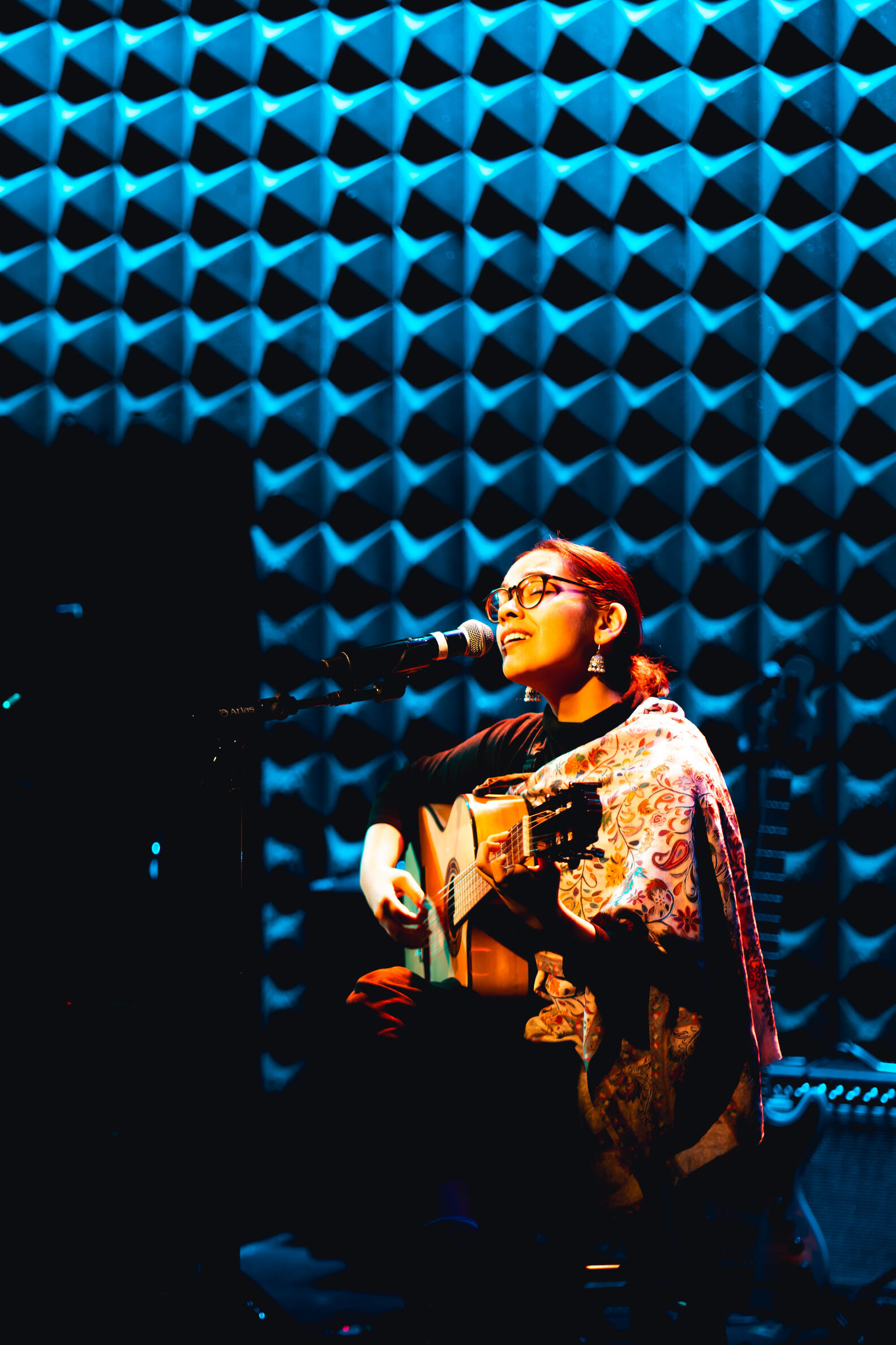 Seated woman with dark hair in a ponytails, wear glasses, playing a guitar