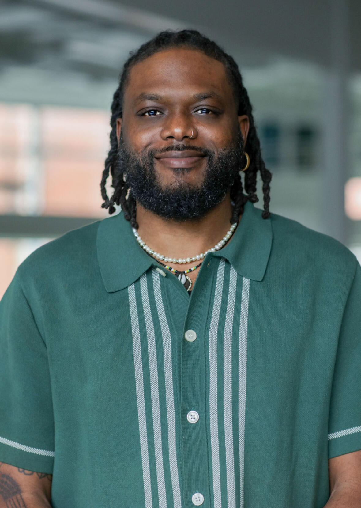 African American man with shoulder length braids and facial hair wearing a green and white shirt, smiling at the camera 