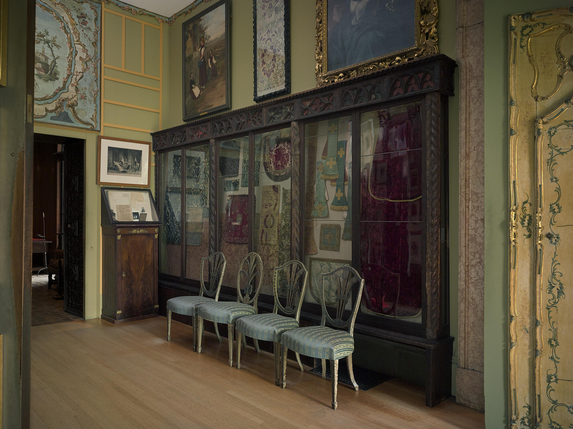 A room with green painted walls and a large wooden and glass case filled with a variety of textiles. Above the case on the left side, hangs Francesc Alexander’s painting of a young woman standing placing flowers in a niche along a road in the Italian countryside.