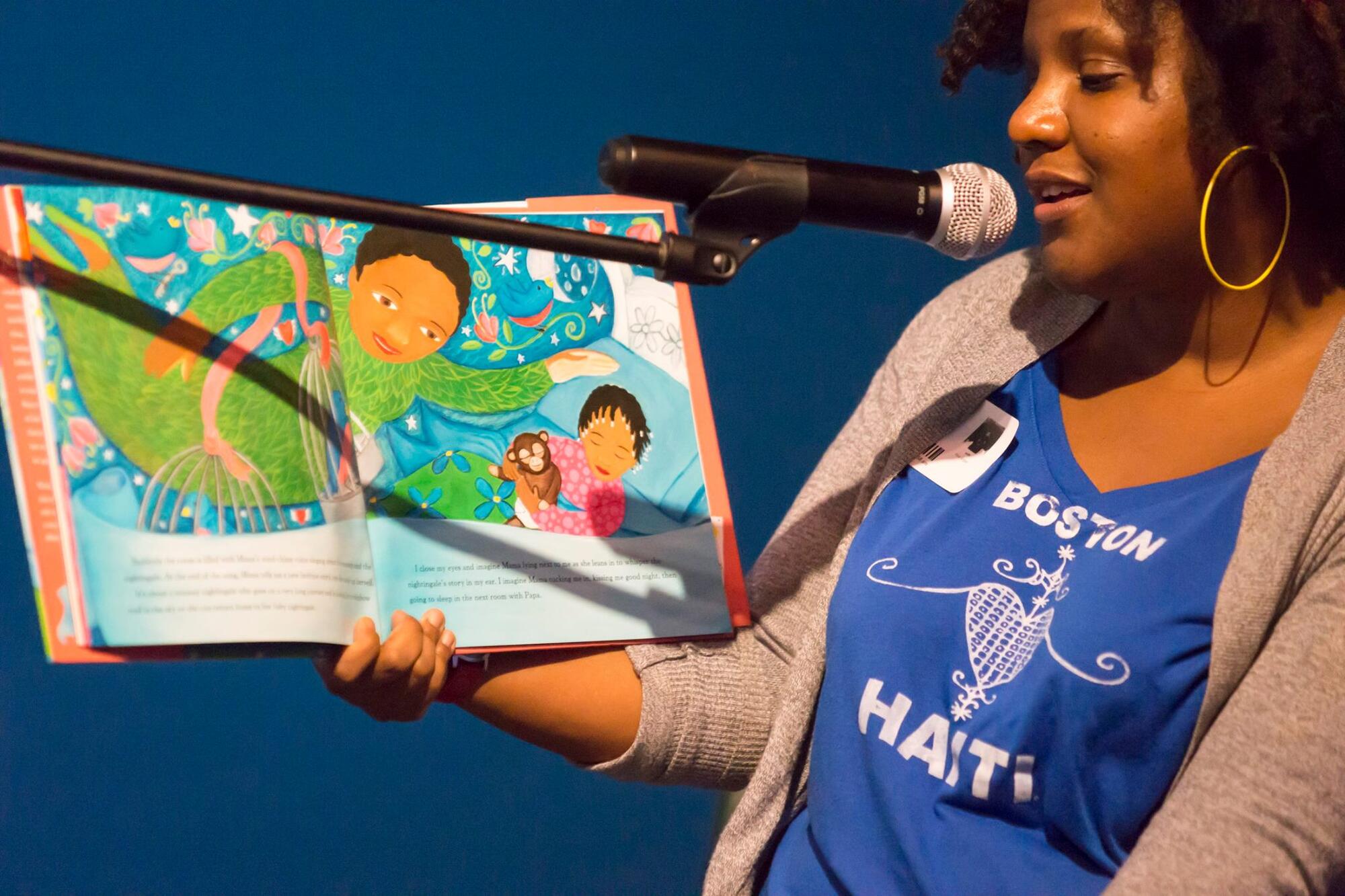 African American woman with large hoop earrings with a blue t-shirt with the words Boston Haitti on the front, reading a children's book in front of a microphone. 